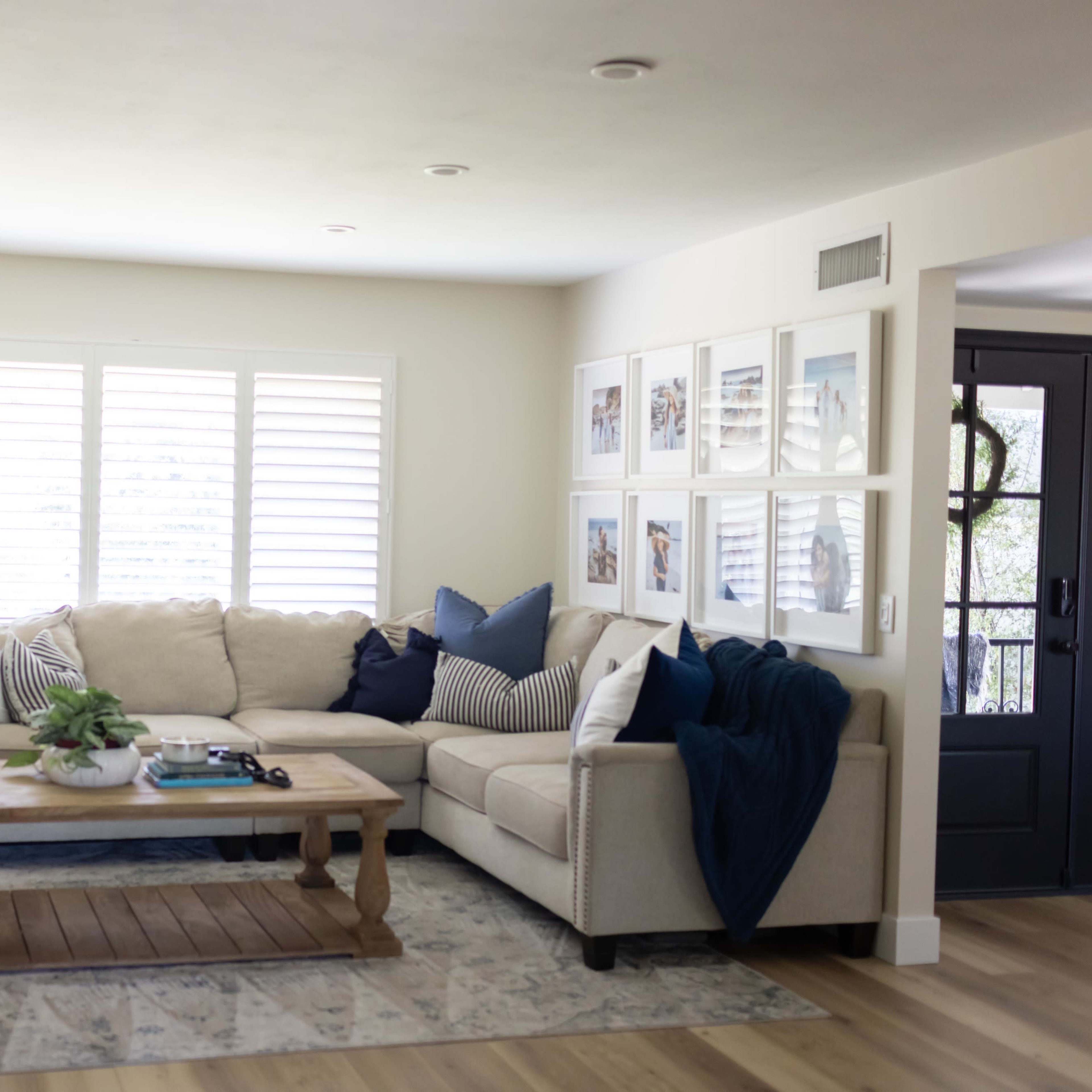 A clean living room with a beige sectional sofa, a wooden coffee table, and framed pictures on the wall beside a set of shuttered windows.
