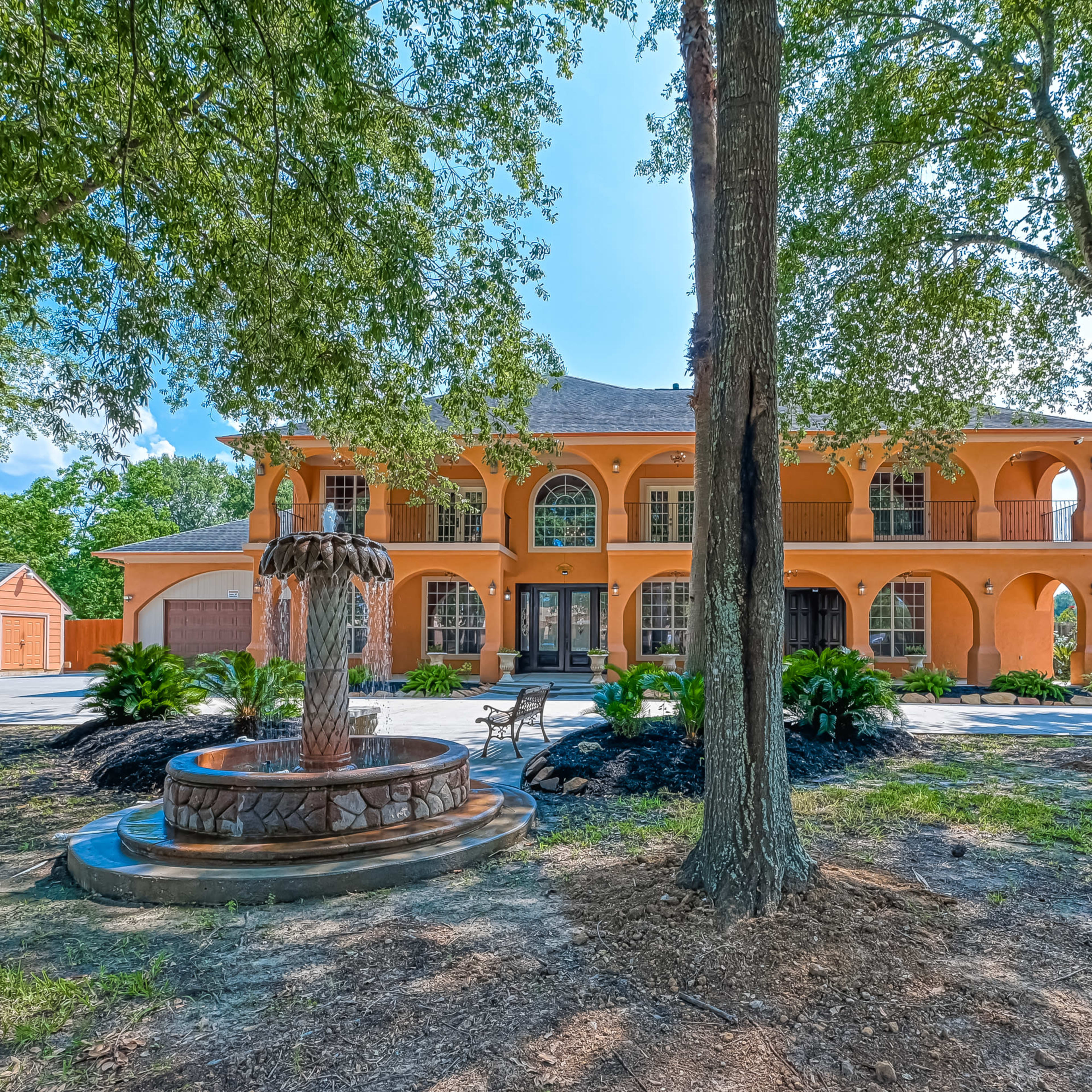 A large orange house with a fountain in the foreground is surrounded by trees and greenery.