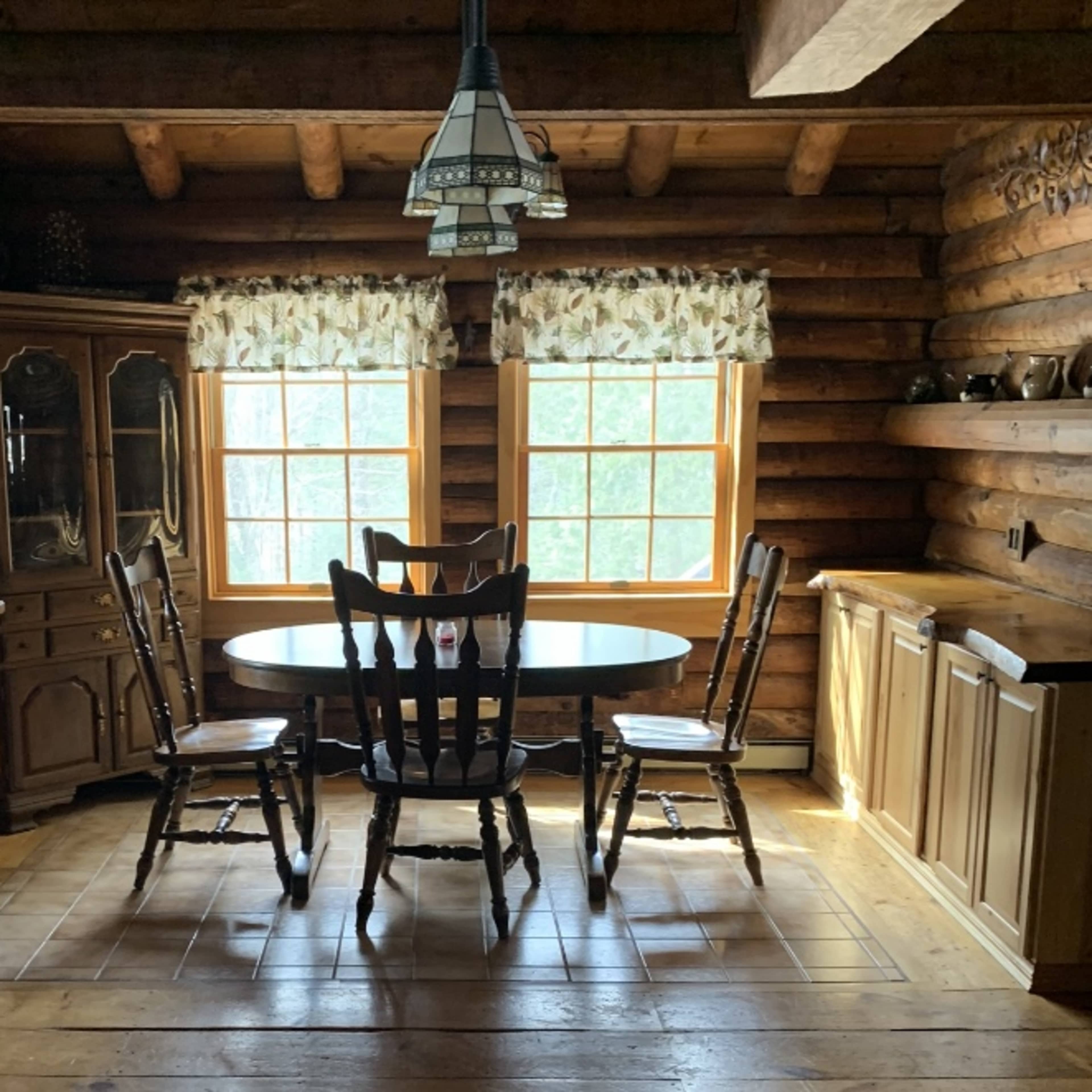 A wooden dining table surrounded by four chairs is situated in a log cabin kitchen with large windows and rustic furniture.
