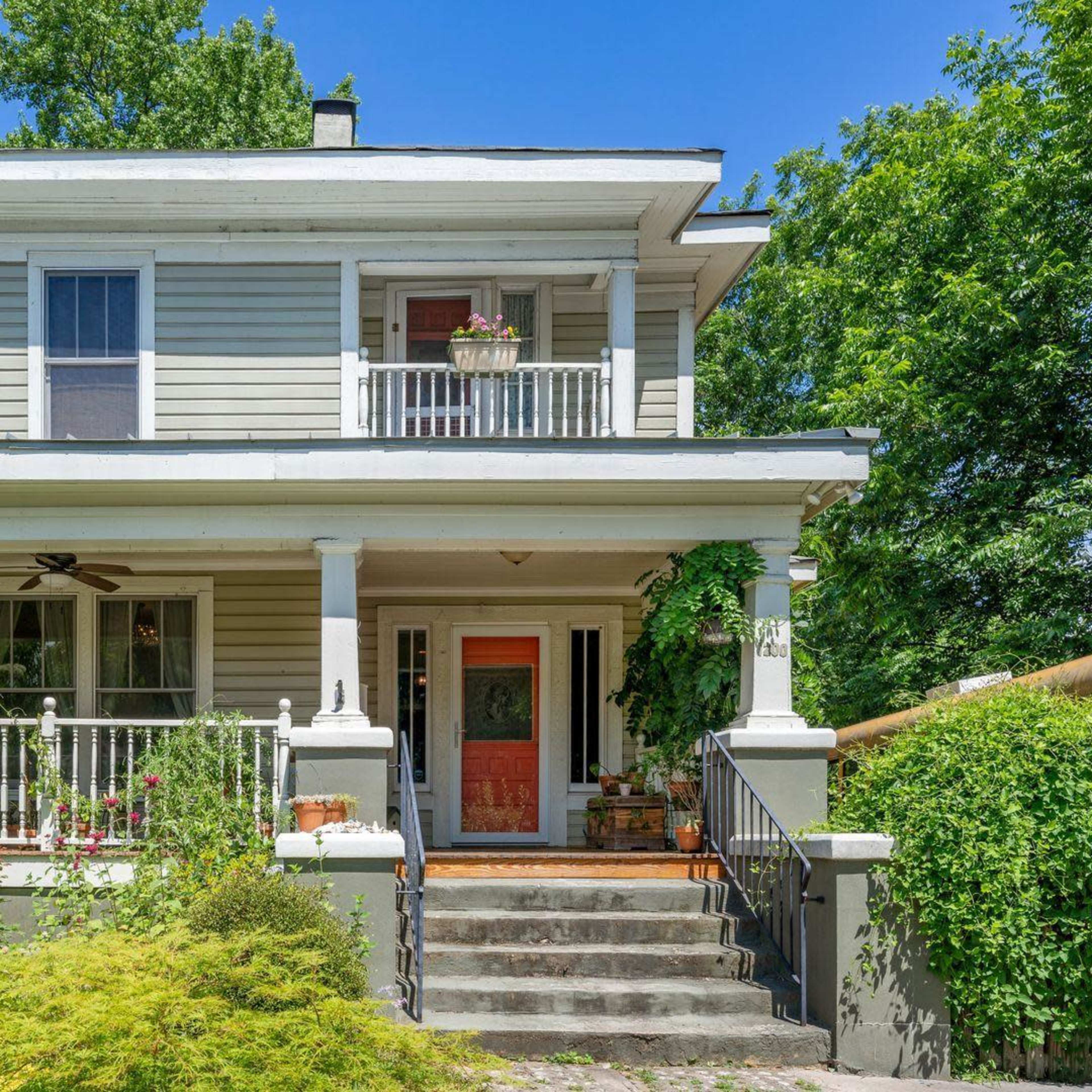 A two-story house with gray siding, a front porch supported by white columns, and lush greenery surrounding the entrance.