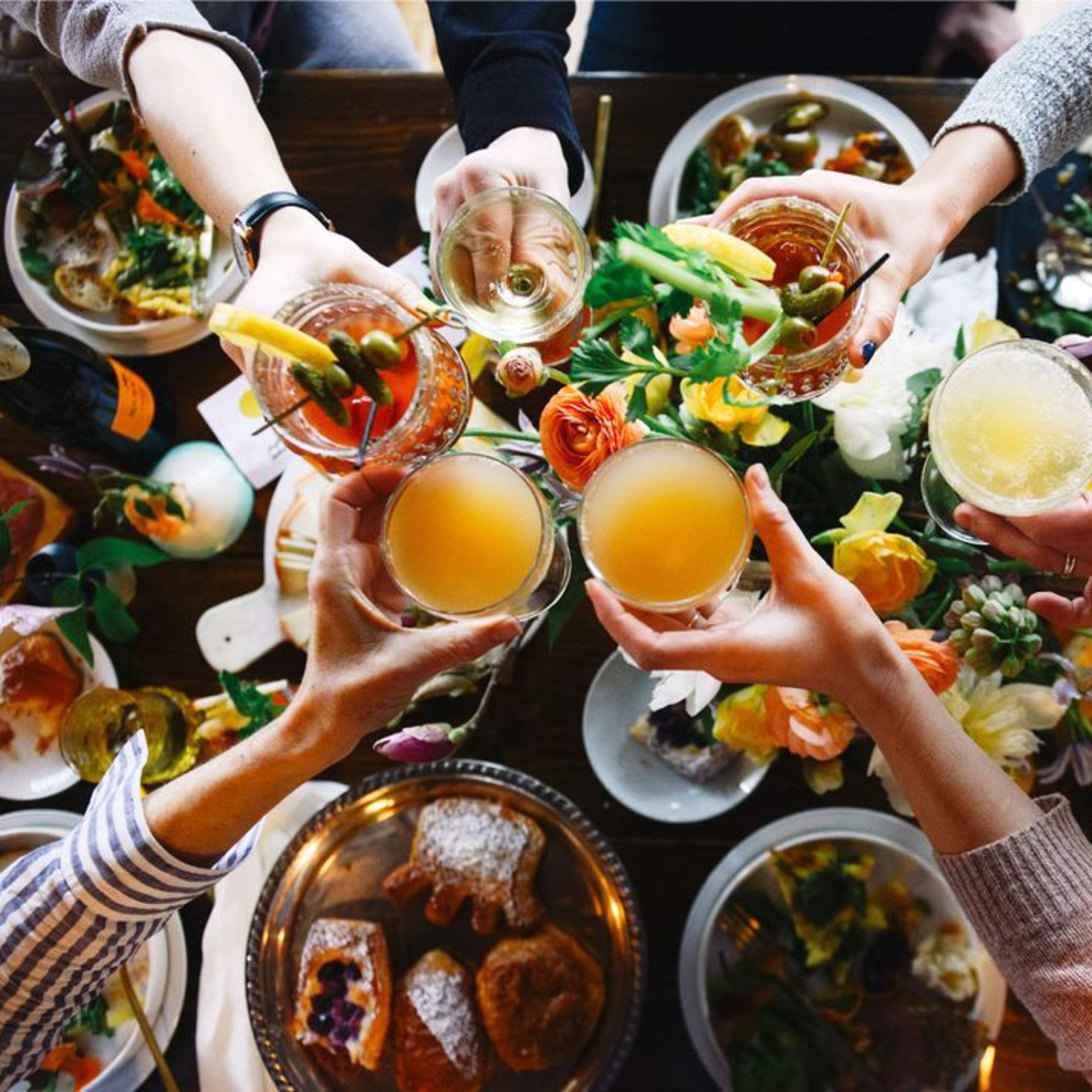 A group of hands raises various drinks in a toast above a table set with food and floral decorations.