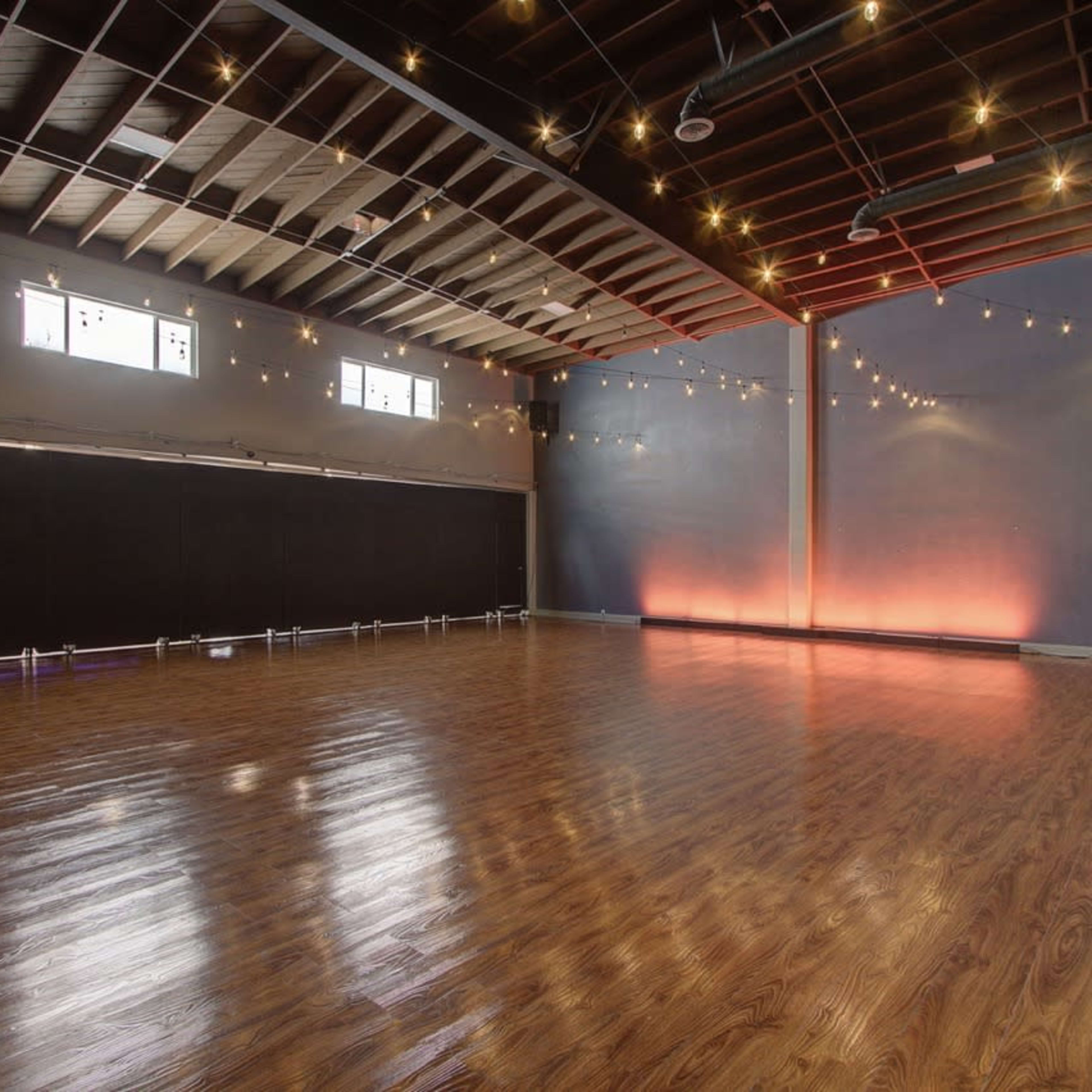 A spacious dance studio features polished wood flooring and soft lighting, with large windows and a black wall at one end.