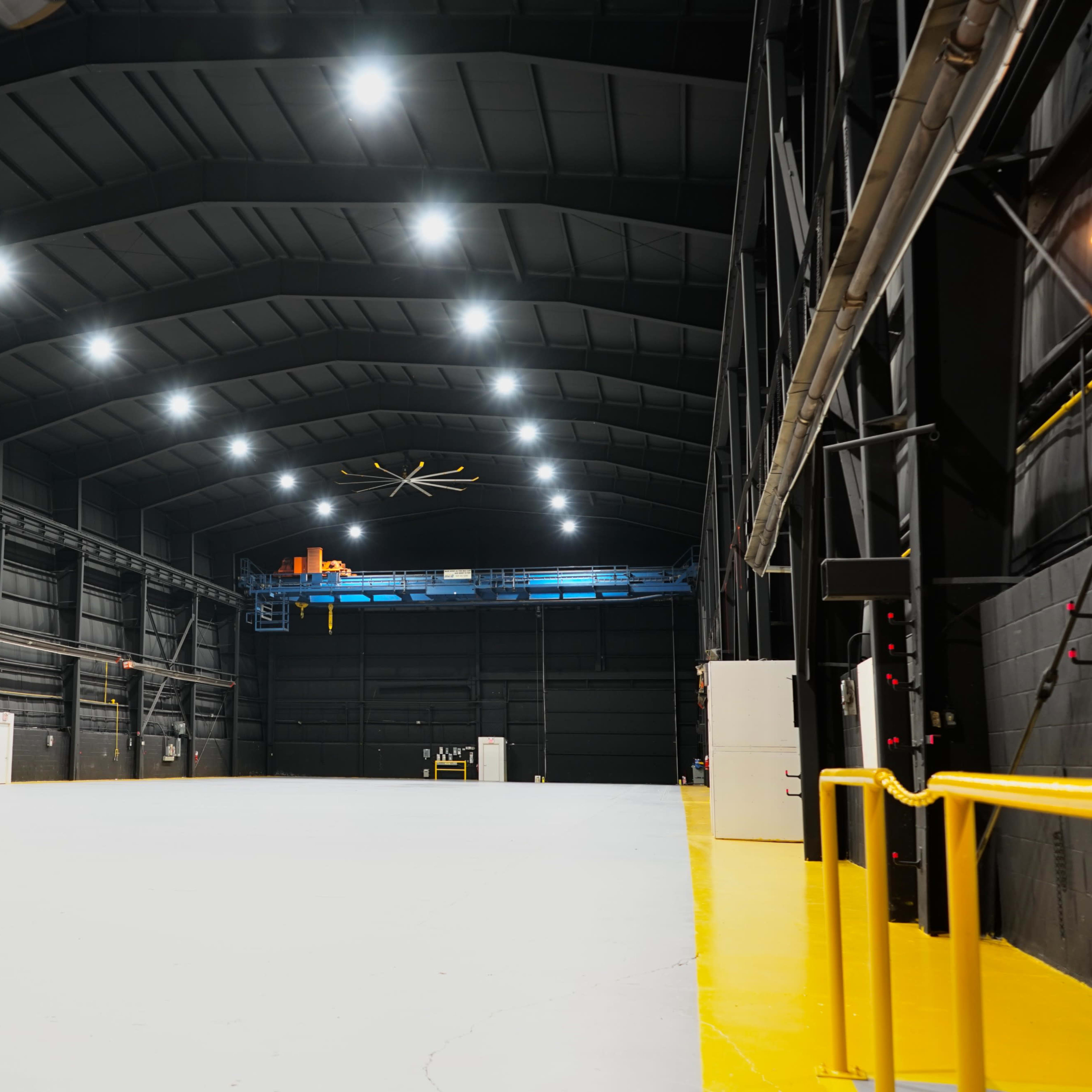 A large, empty industrial warehouse with a high ceiling, bright overhead lights, and a wide, open floor space.