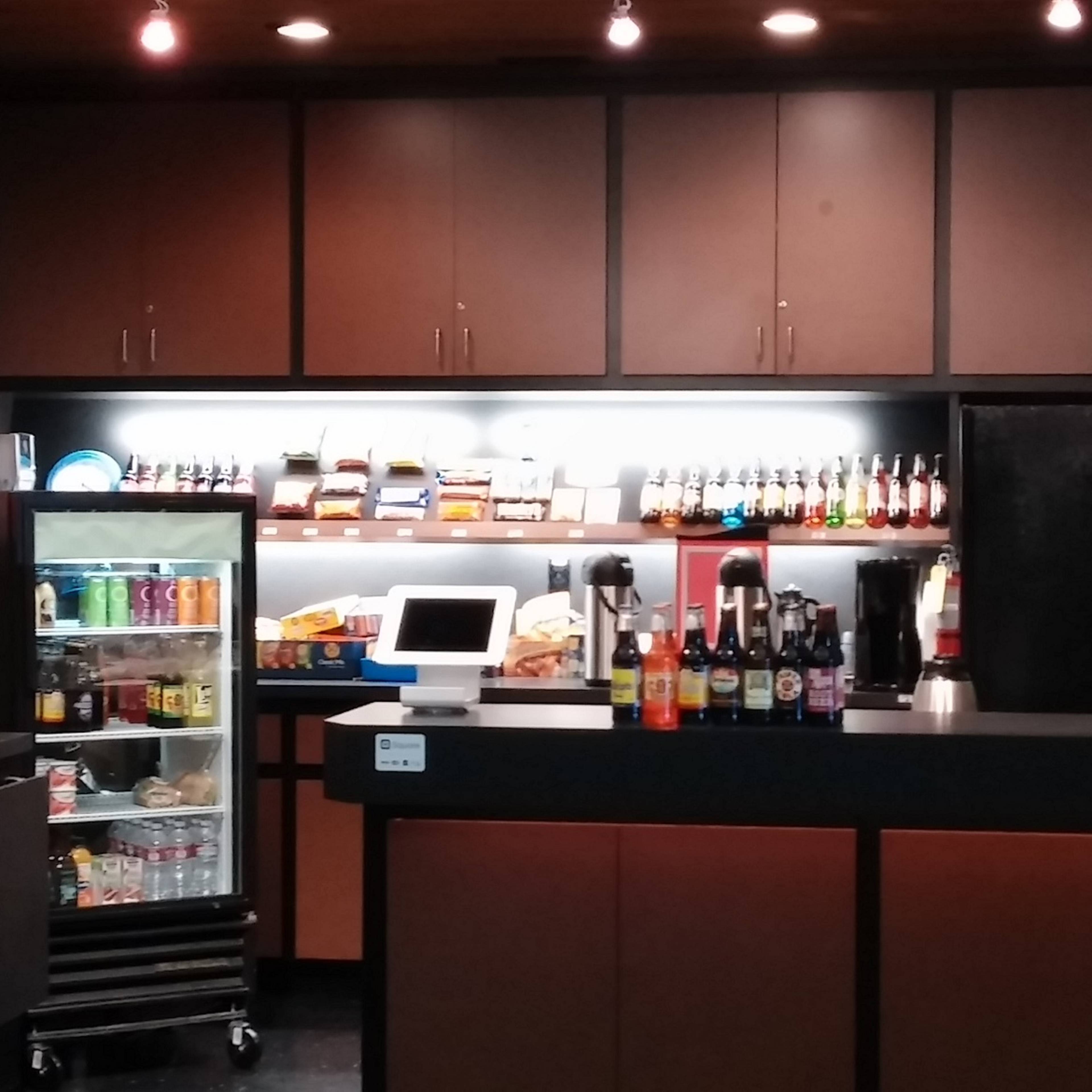 A concession stand with a black counter, displaying various beverages and snacks under bright lighting, along with a fridge filled with drinks and several shelves of food items above.
