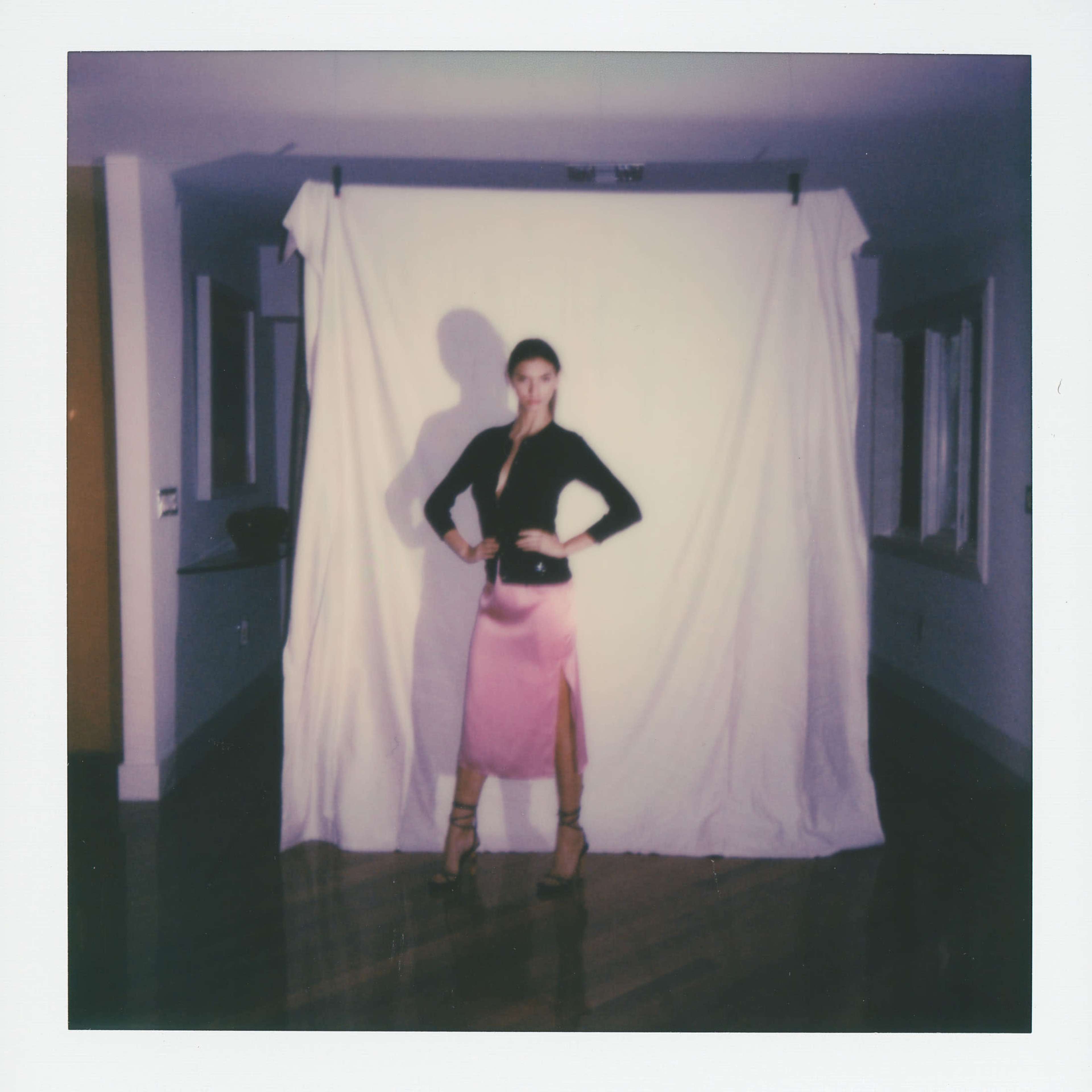 A person stands posing in front of a white backdrop in a room with wood flooring.