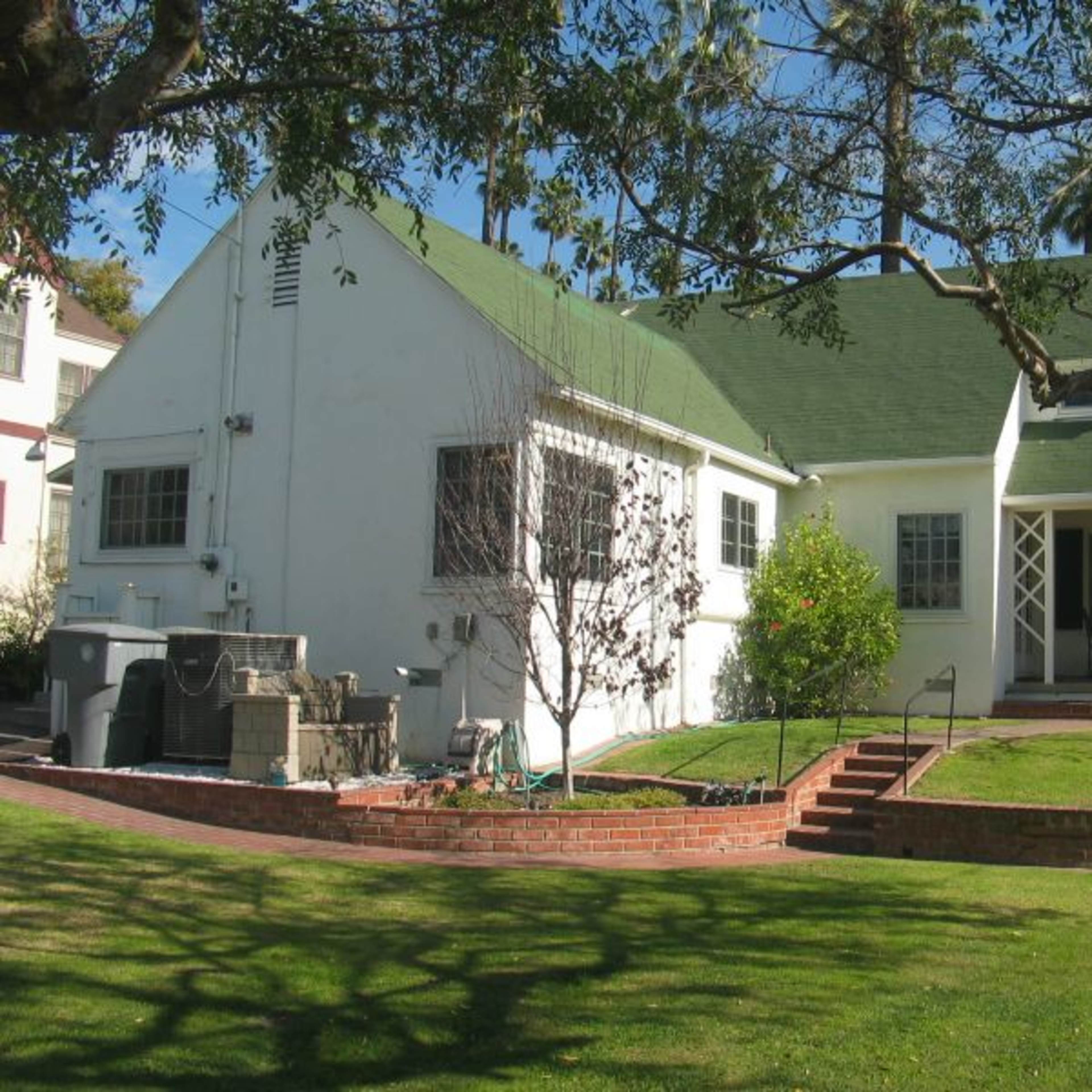 A white house with a green roof, accompanied by a landscaped yard featuring a small patio and several trees.