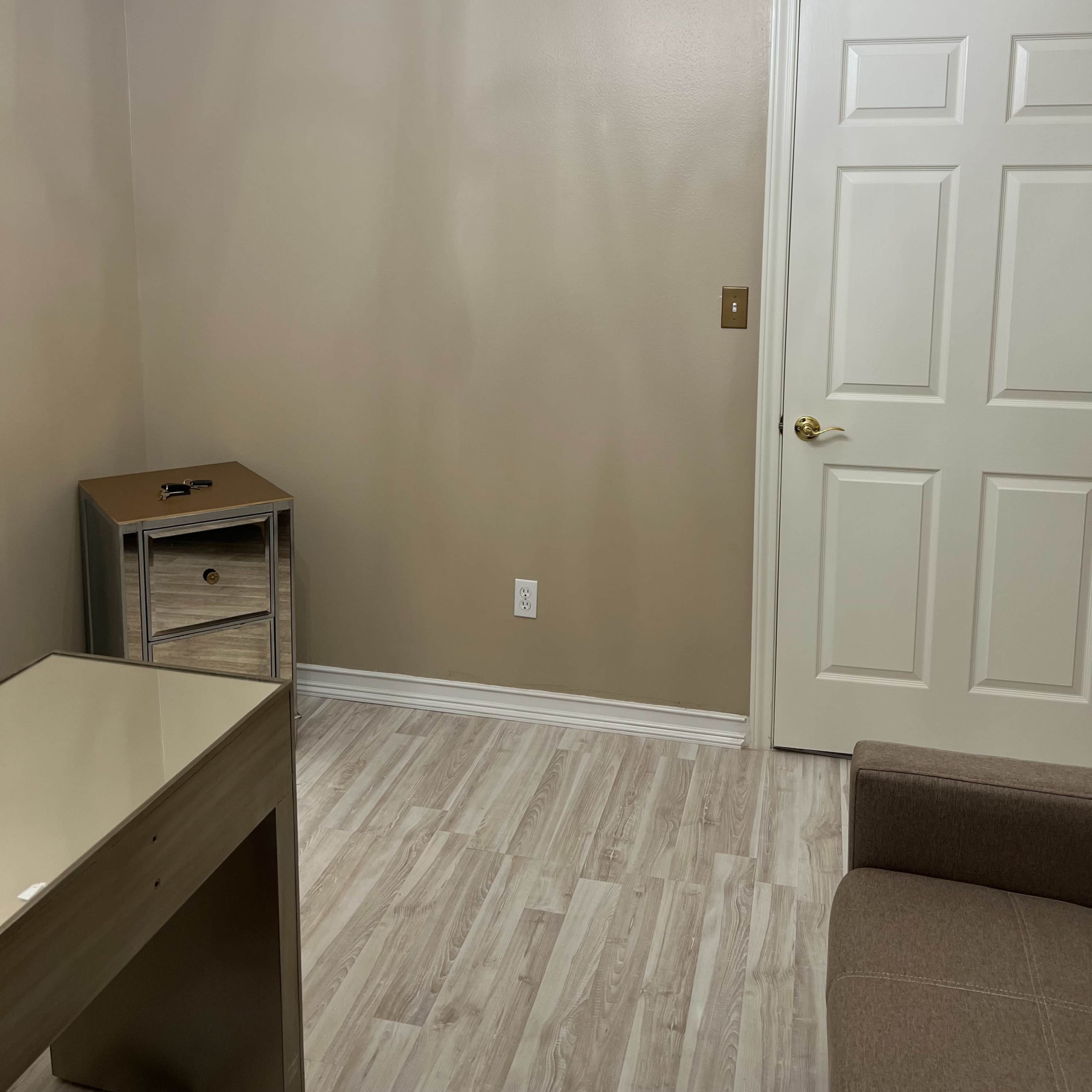 A small room with a chandelier, beige walls, a light-colored floor, a gray couch, a cabinet with a drawer, and a closed white door.