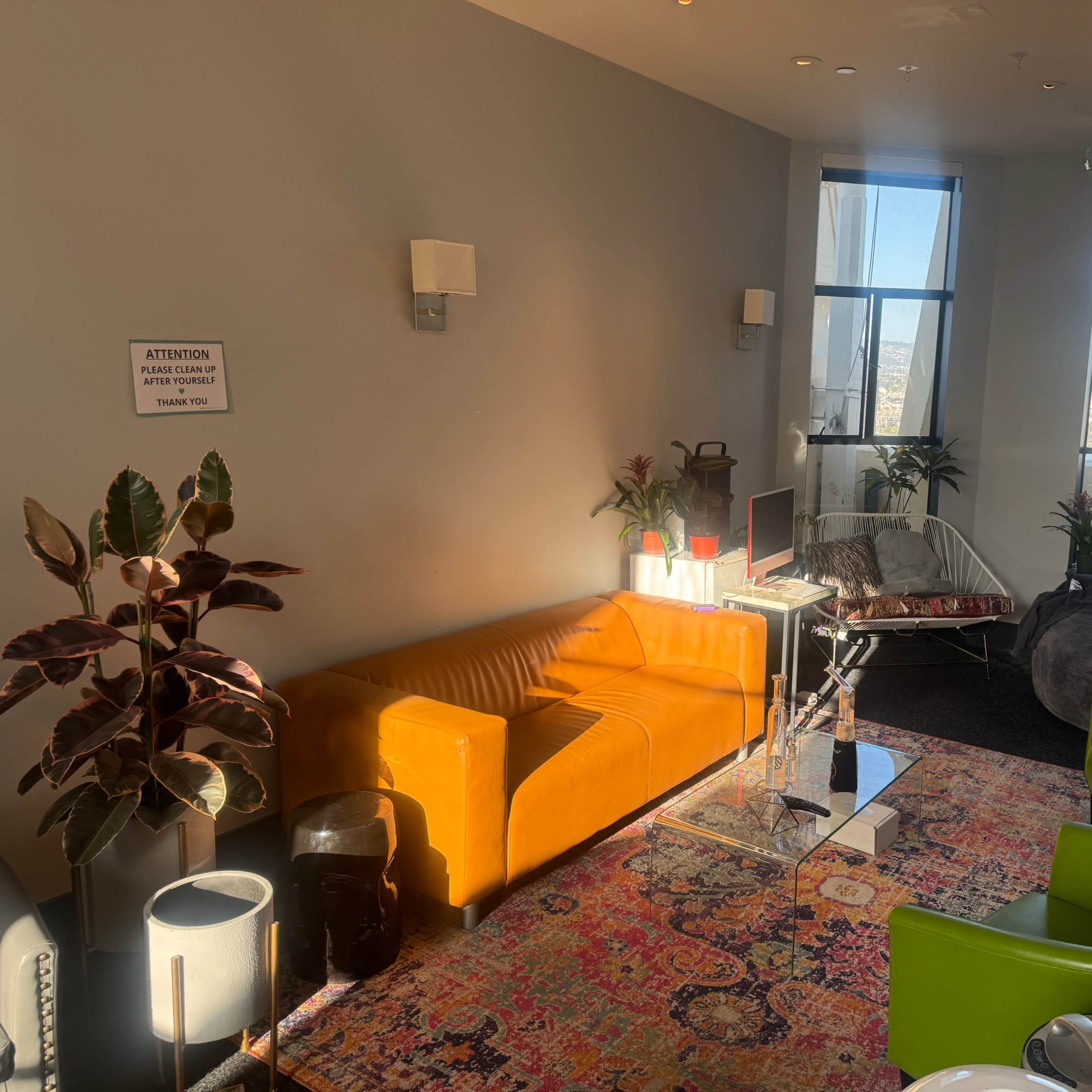 A bright room features an orange sofa, two green chairs, and a table with plants, along with a view of the city through large windows.