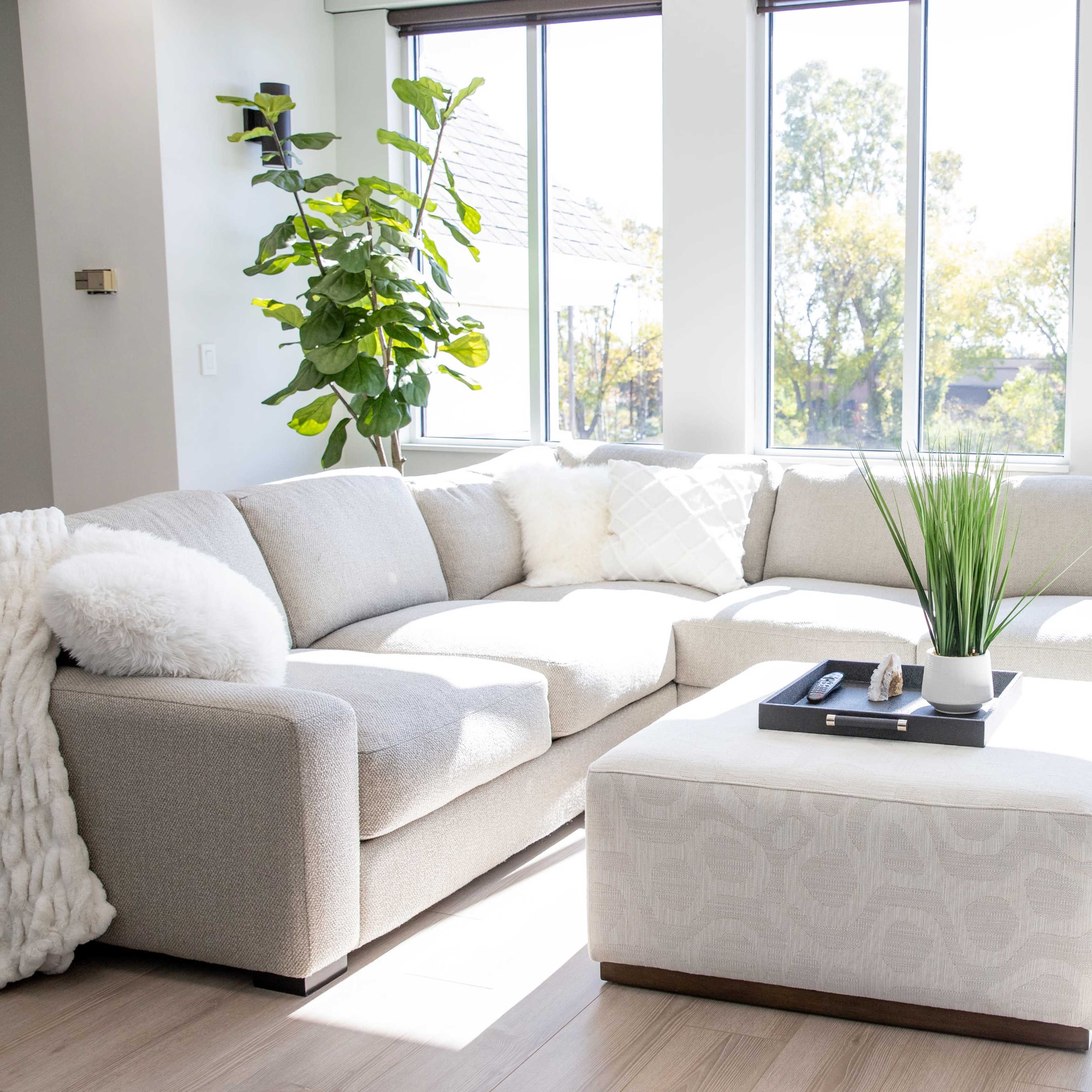 A spacious living room features a light gray sectional sofa and a matching ottoman, alongside large windows that allow natural light to illuminate the space.