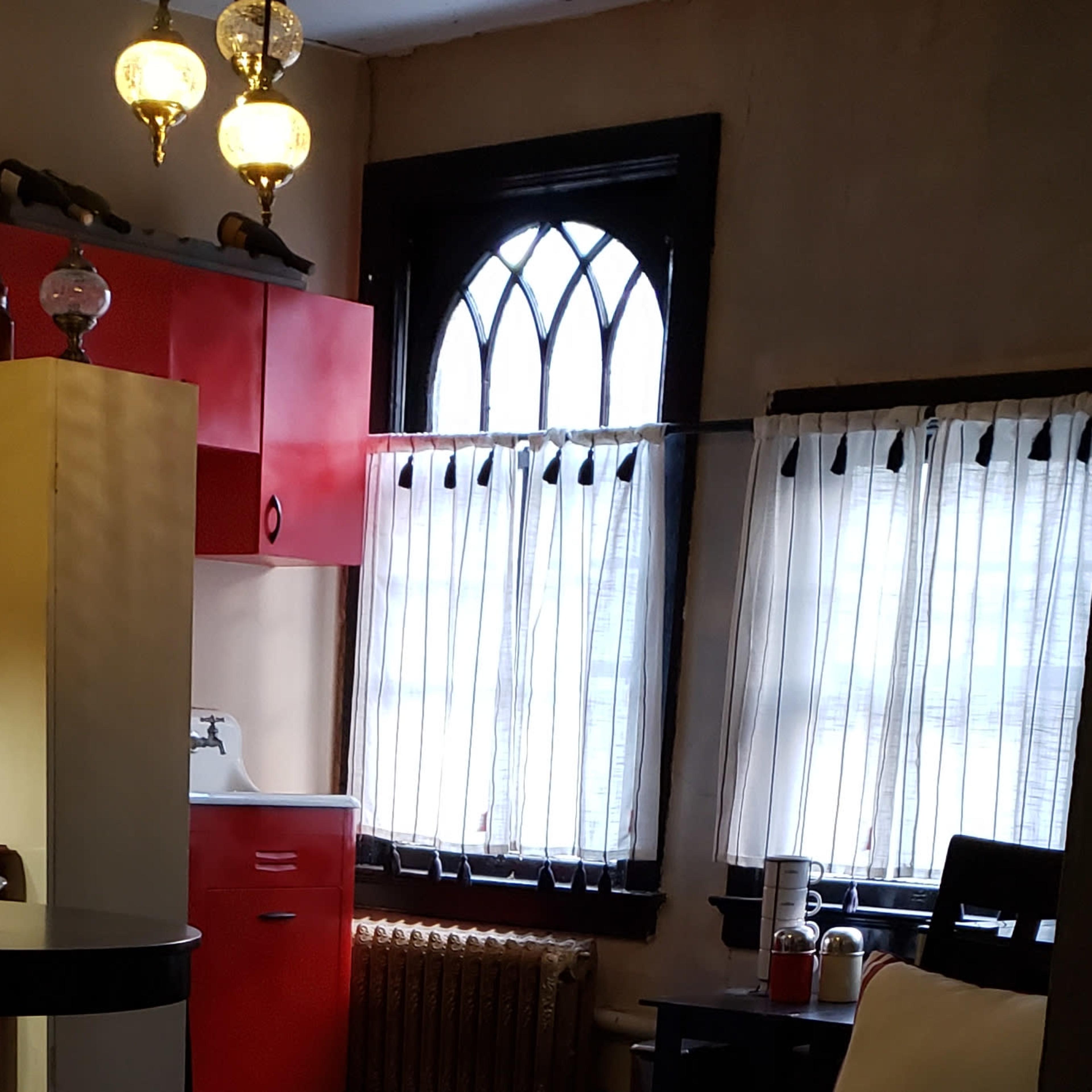 A kitchen area with red cabinetry, a white sink, and a window adorned with sheer curtains, set in a room with patterned walls and vintage lighting fixtures.