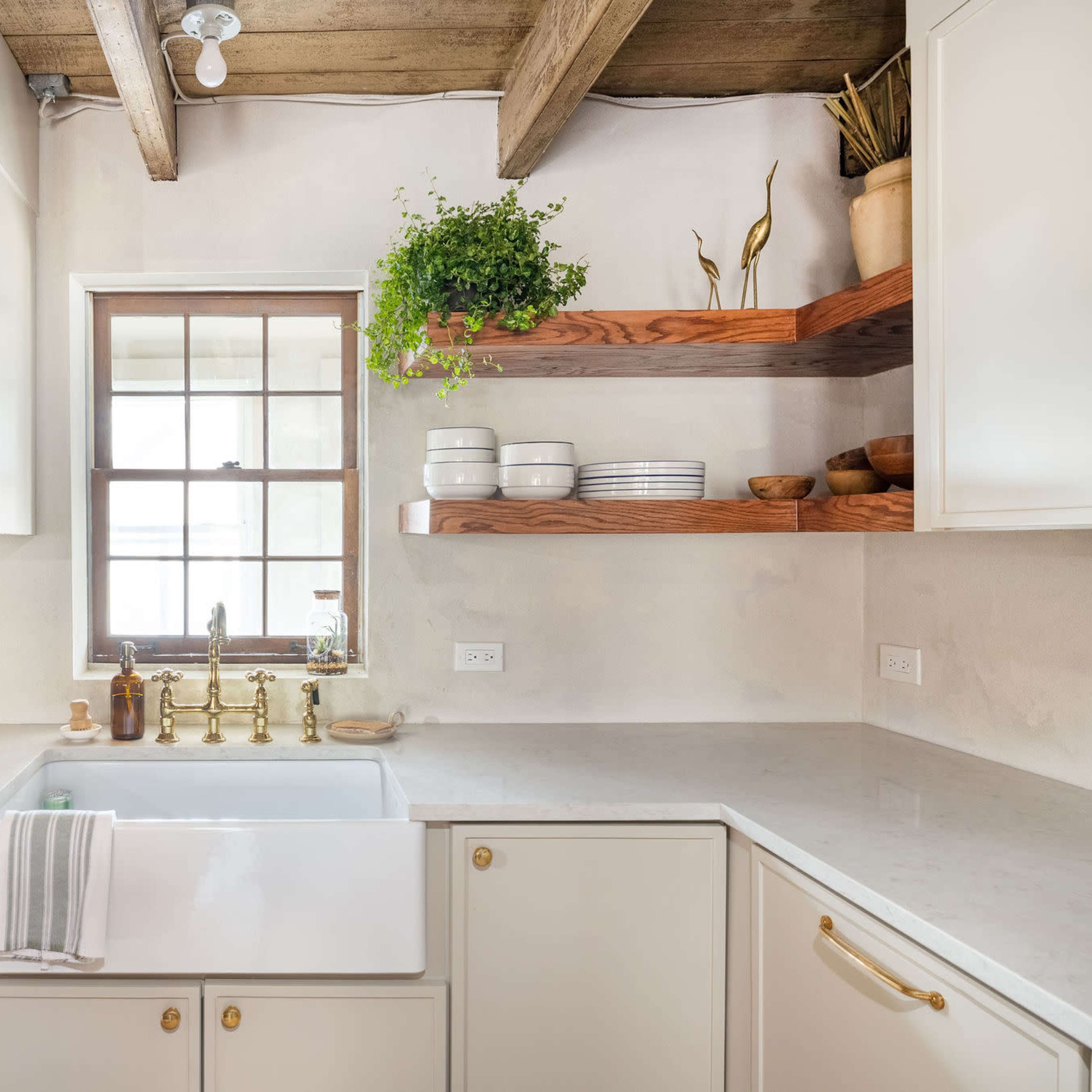 A bright kitchen features white cabinetry, a farmhouse sink, open wooden shelves, and brass fixtures.