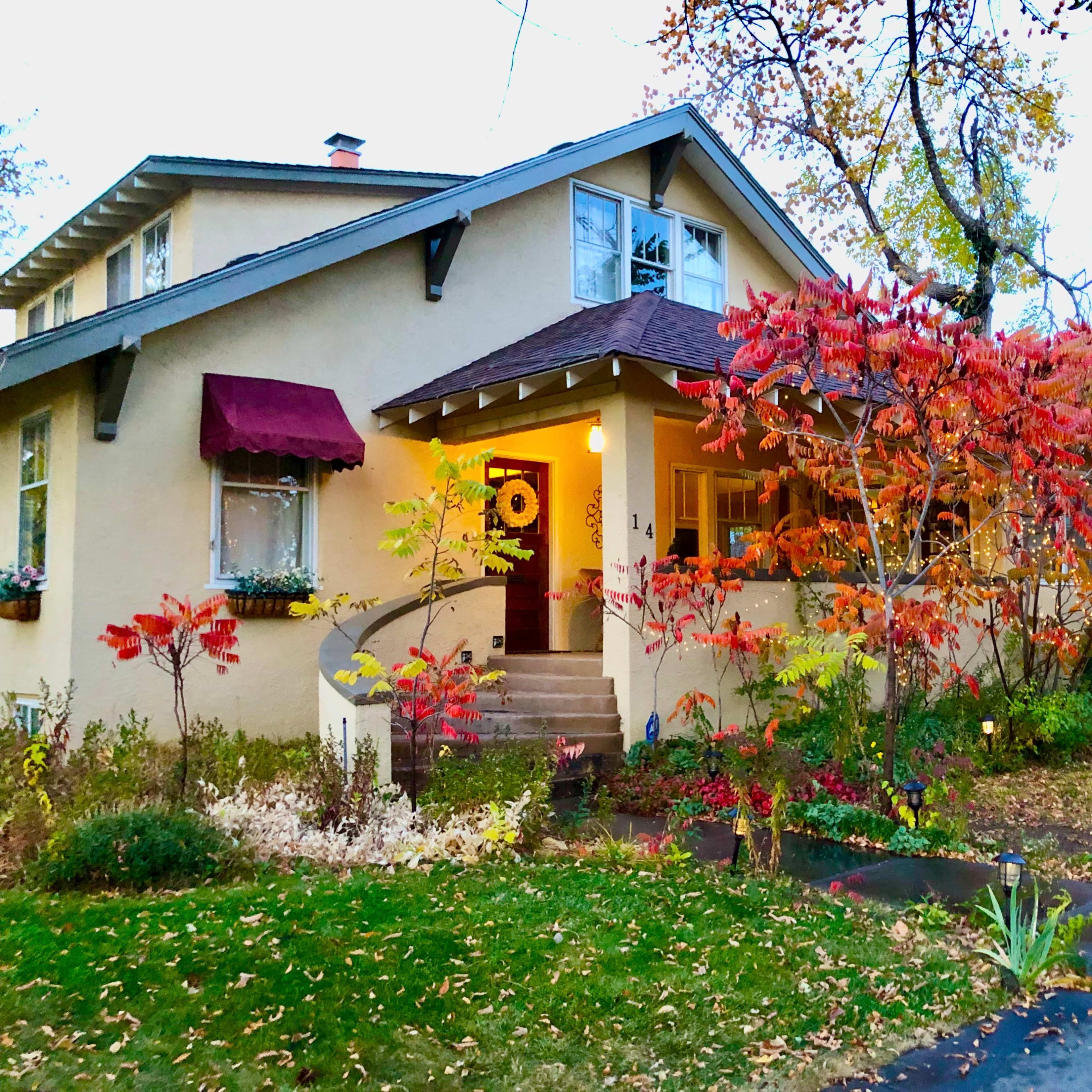 A two-story house with a maroon awning features colorful autumn foliage in the front yard and a well-kept garden.