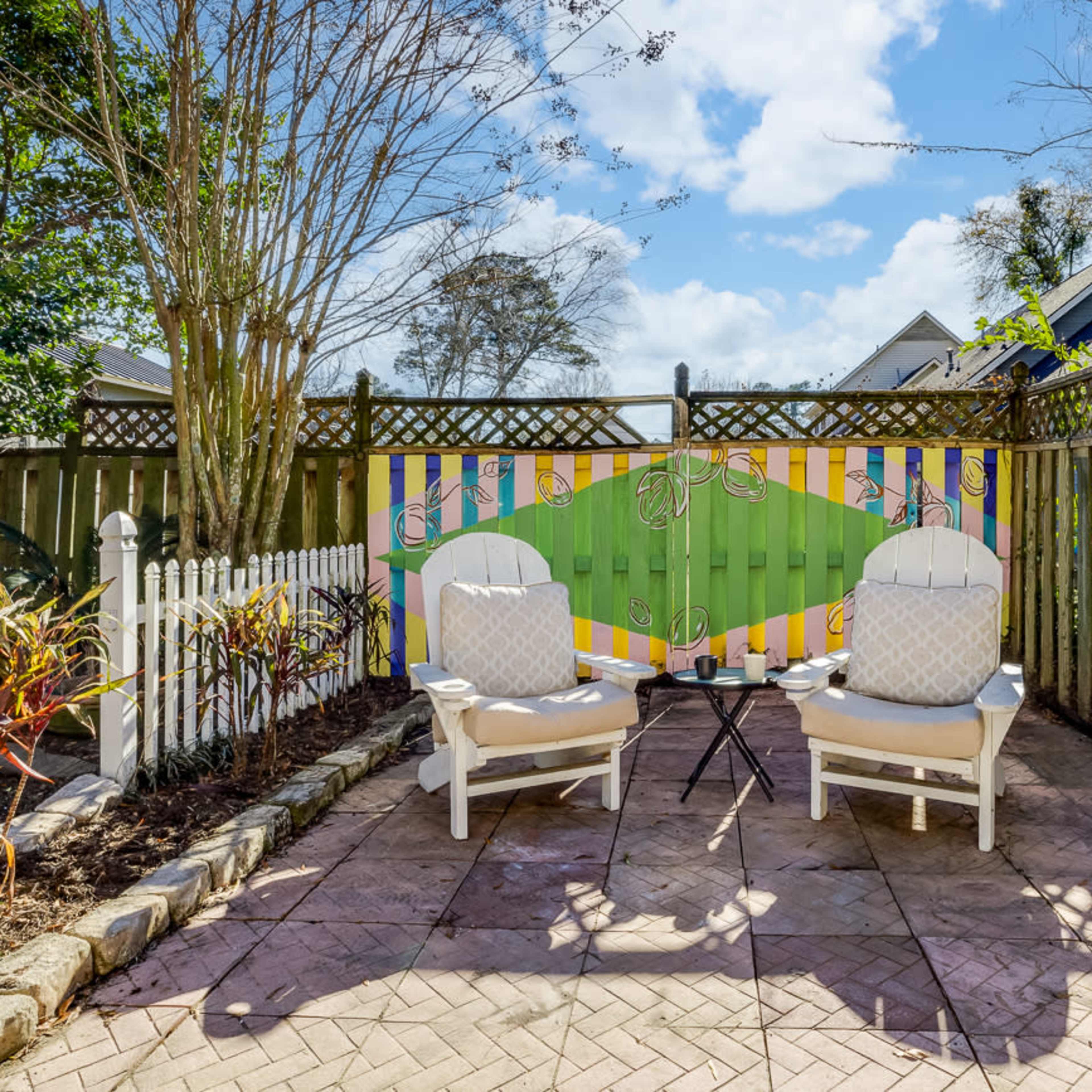 A small, enclosed patio features two white chairs facing a colorful painted wall, surrounded by trees and a garden.