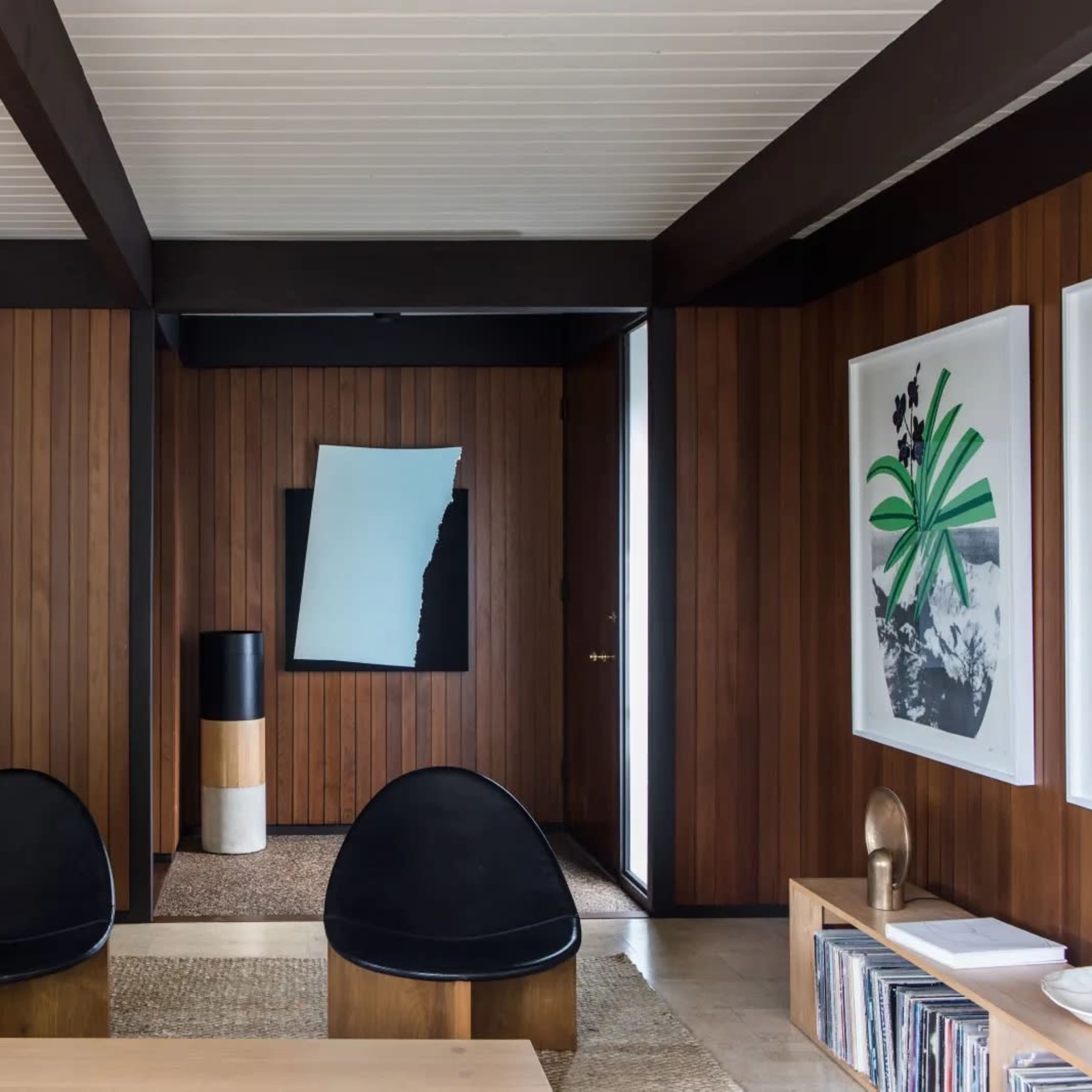 The interior features a modern living space with wooden paneling, two black chairs, a minimalist floor lamp, and art displayed on the walls.