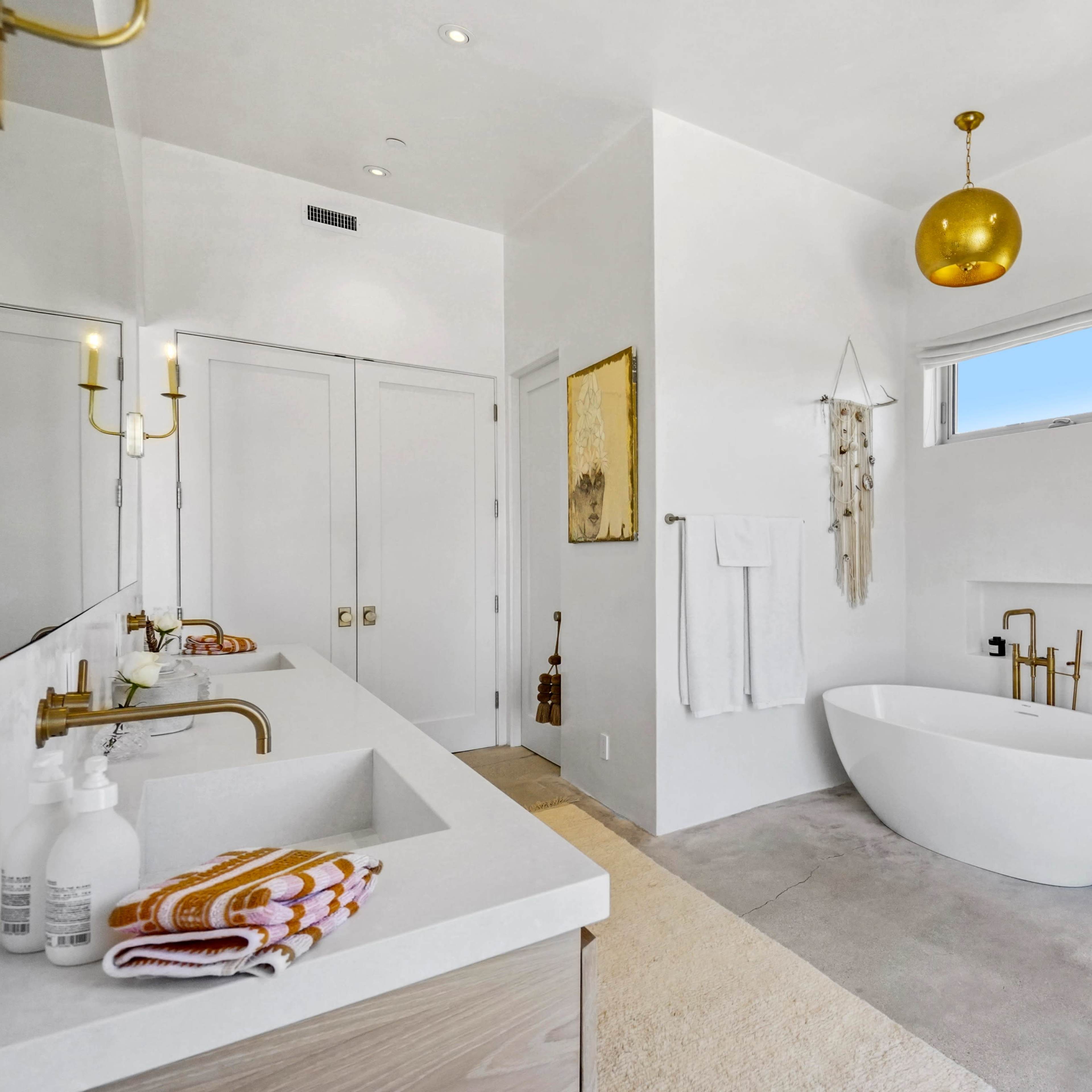 A modern bathroom features a freestanding bathtub, dual sinks with gold fixtures, and large windows allowing natural light.