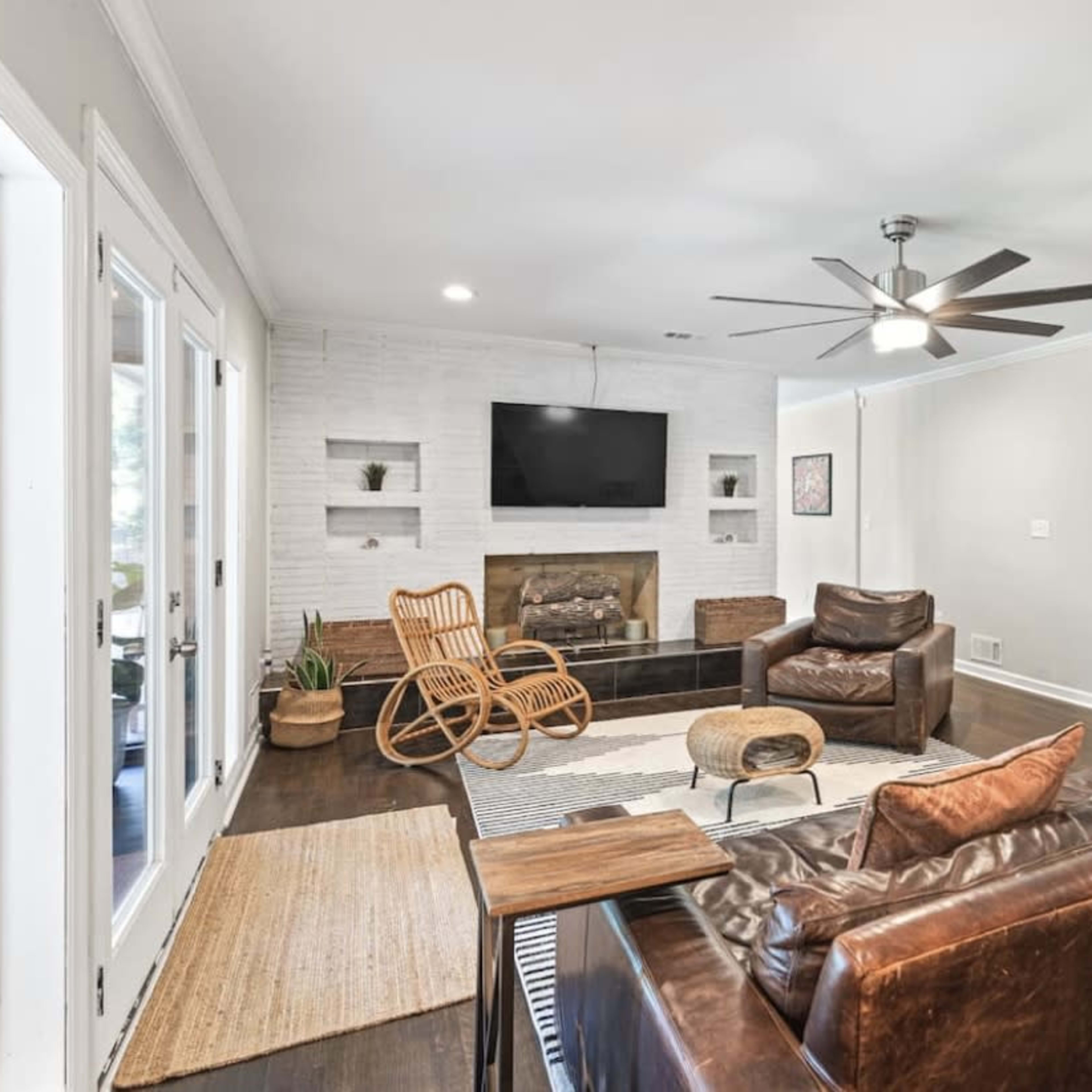 A modern living room with a brown leather sofa, a matching armchair, a wooden rocking chair, a wall-mounted television, and a light-colored brick fireplace.
