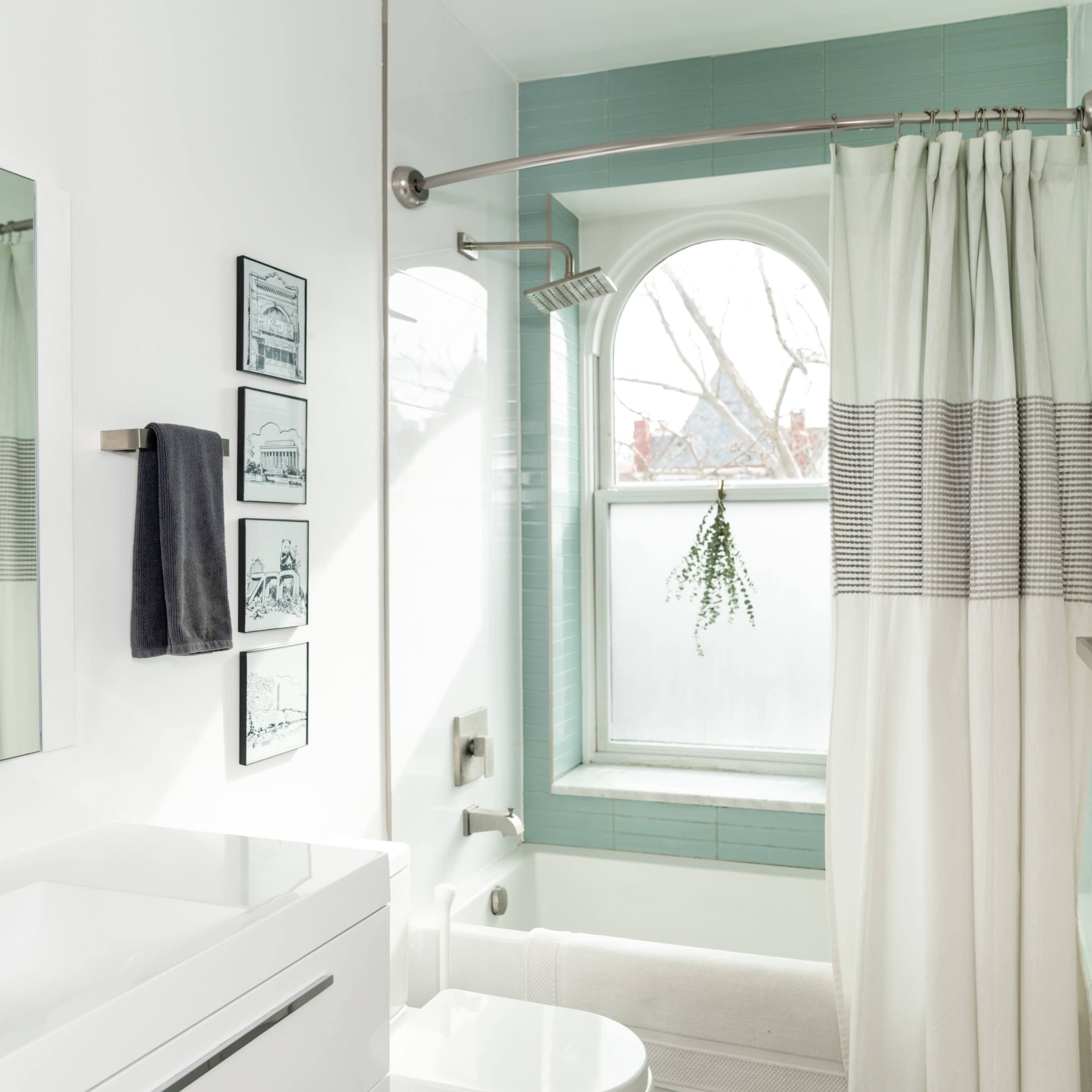 A modern bathroom featuring a bathtub, shower curtain, sink with a mirror, and a window with a plant hanging inside.
