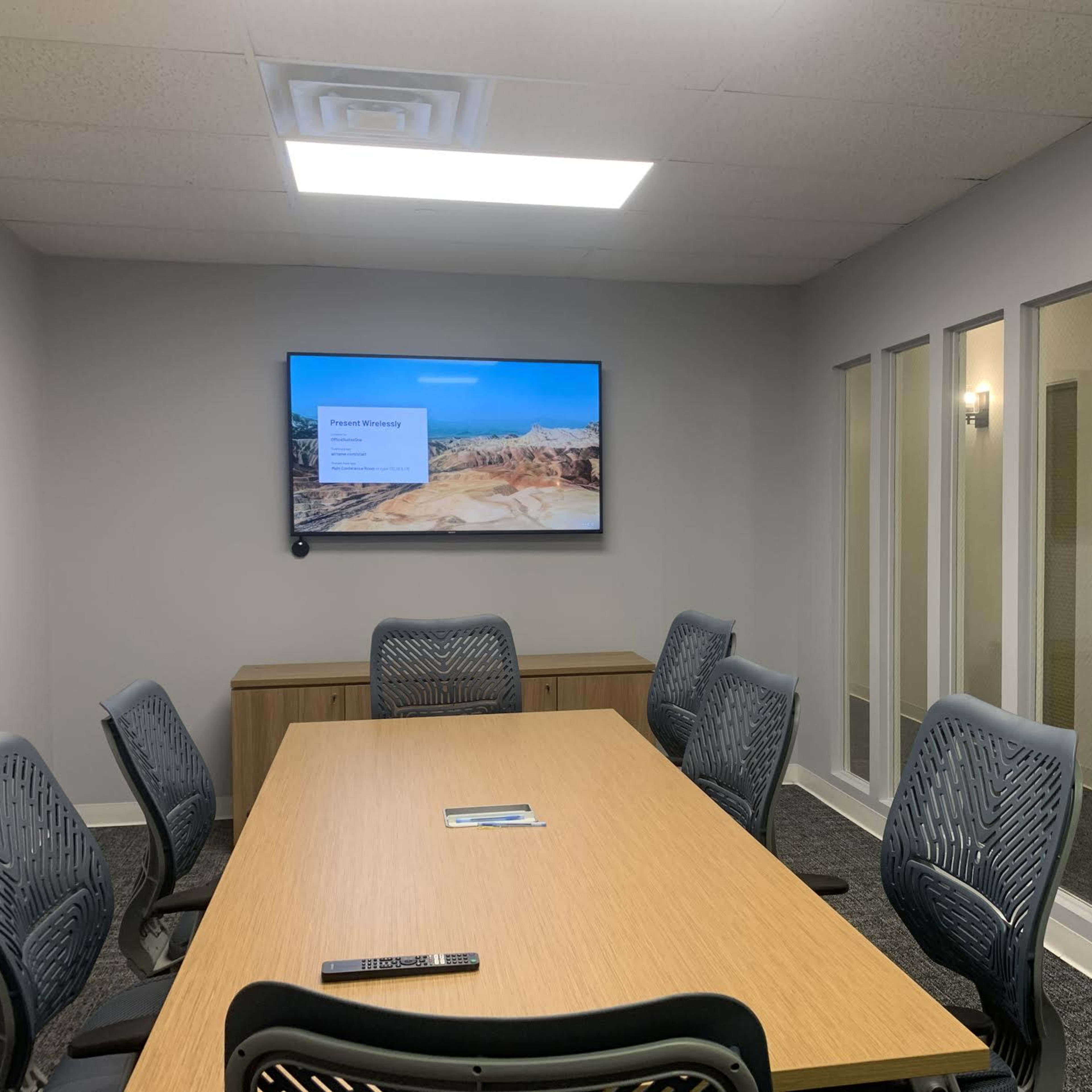 A conference room features a large wooden table surrounded by ergonomic chairs, with a mounted screen displaying a presentation and glass-paneled walls separating it from an adjacent space.