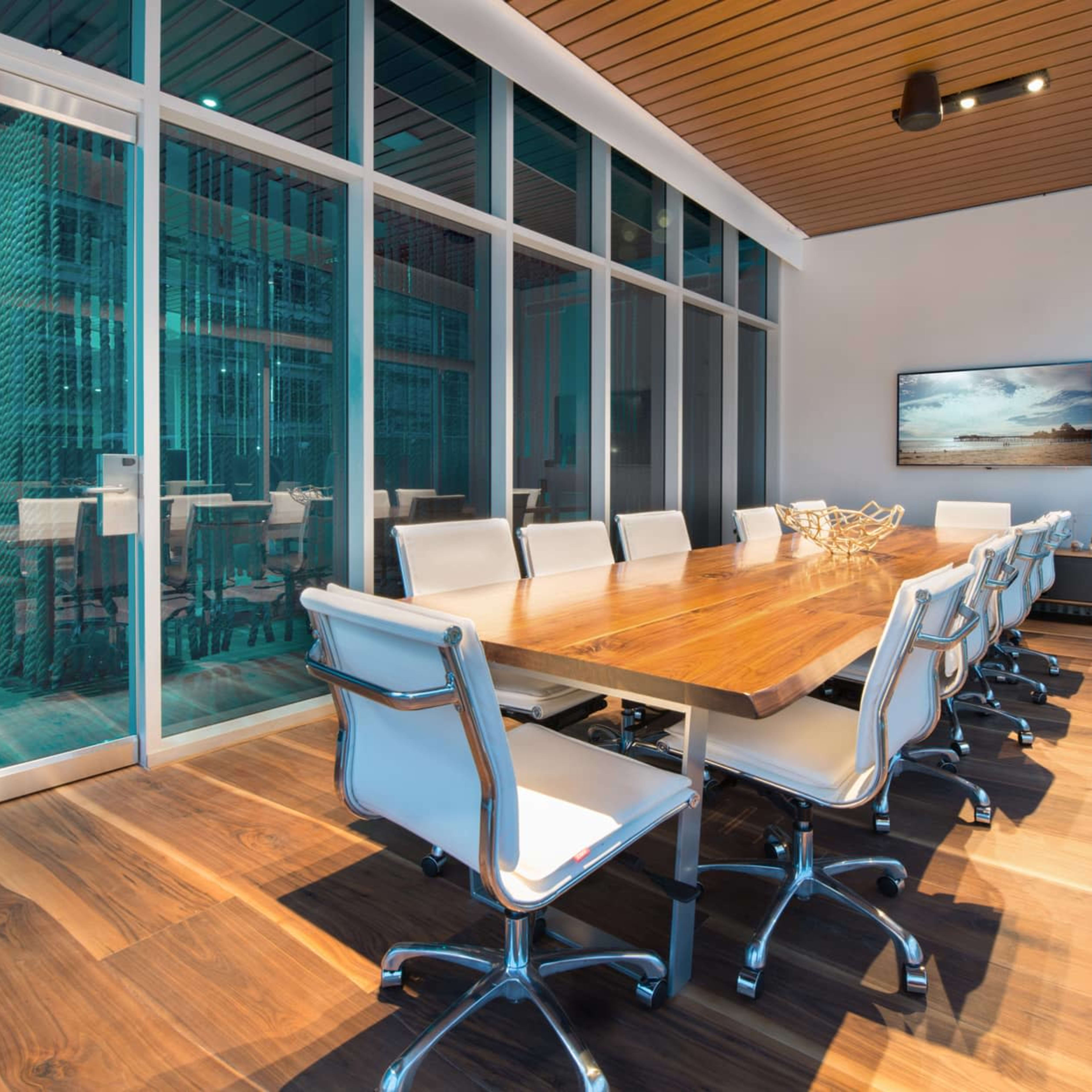 A modern conference room features a long wooden table surrounded by white chairs, with large windows allowing for ample natural light.
