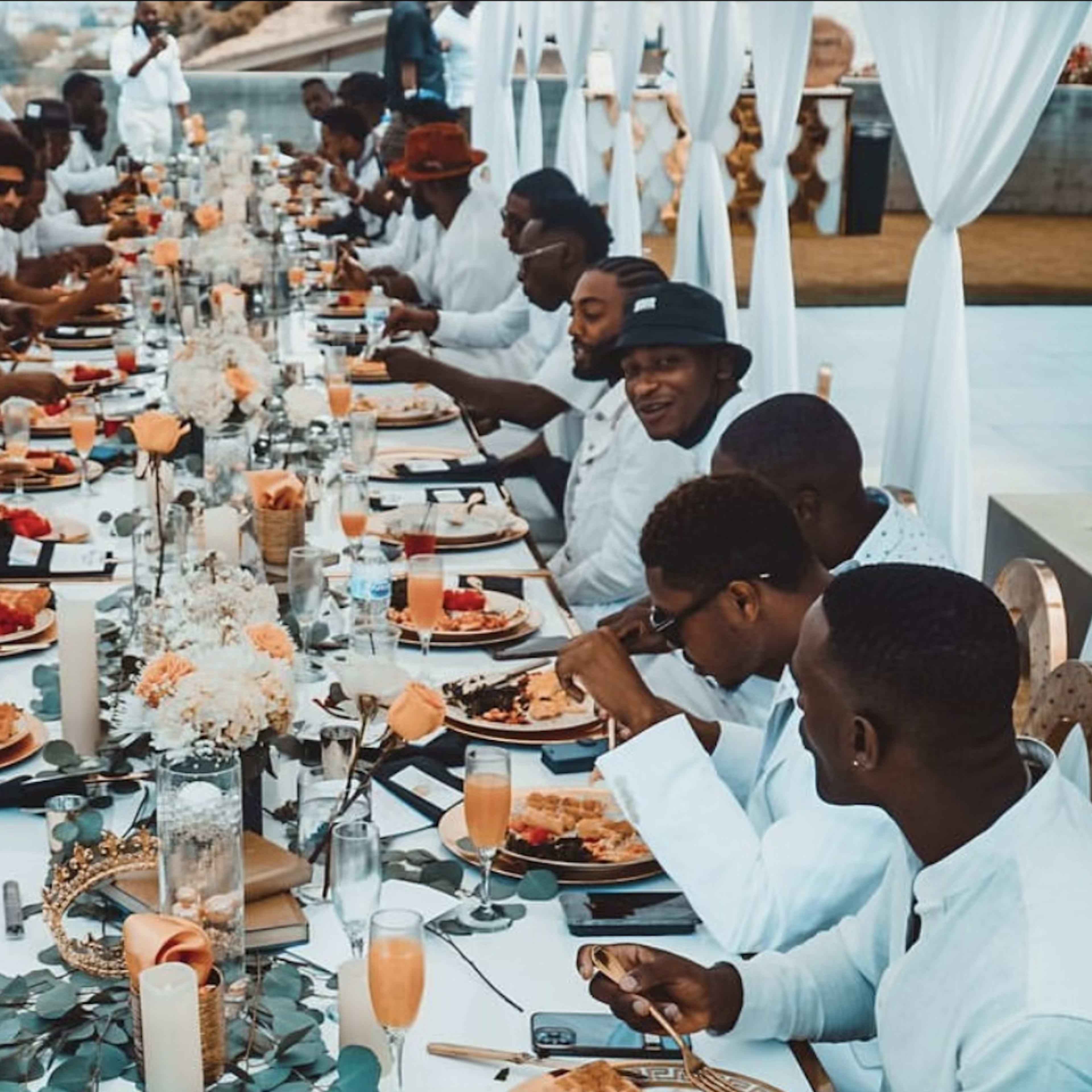 A long banquet table is set outdoors with numerous men in white shirts enjoying a meal, surrounded by elegant decorations and floral arrangements.