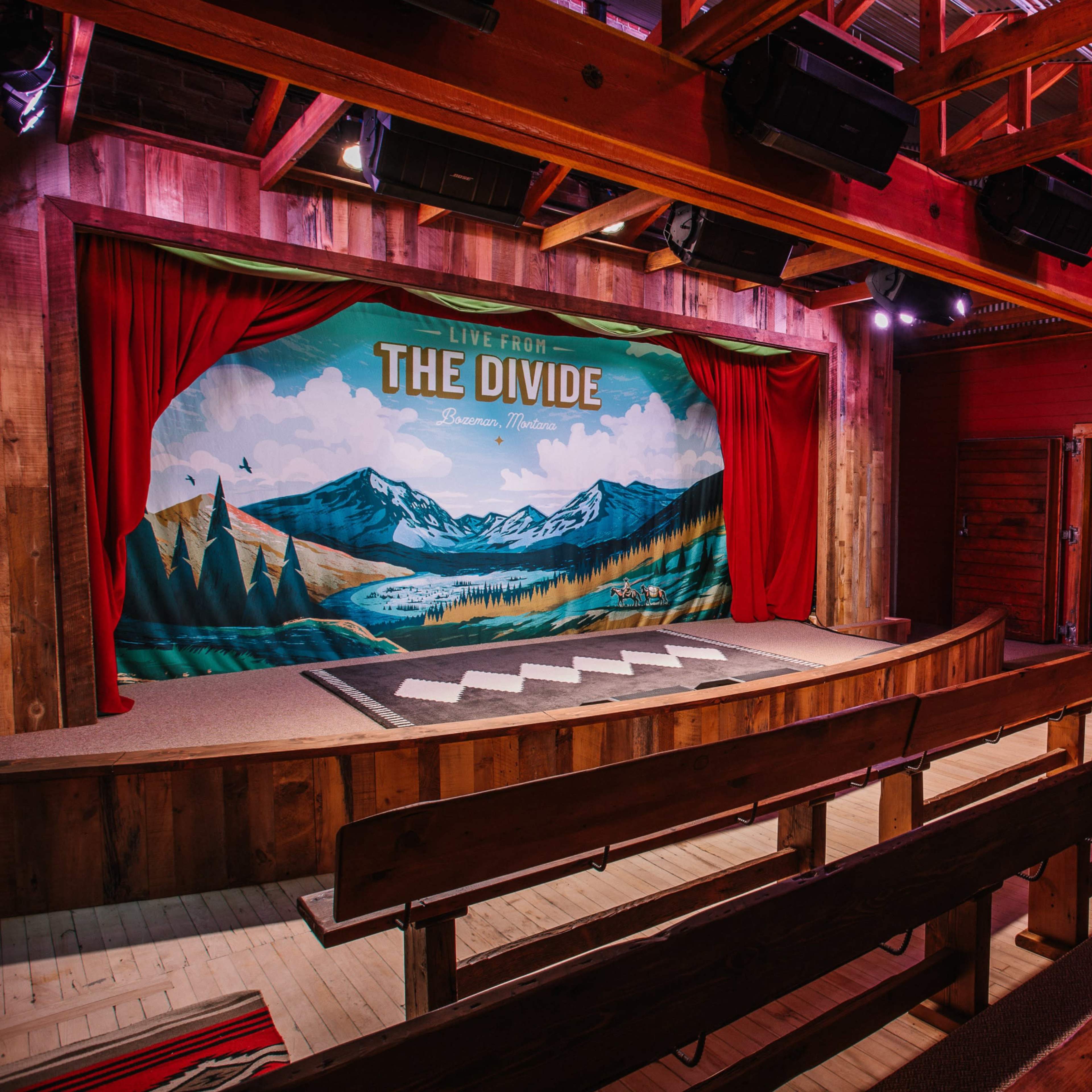 A wooden stage with a painted backdrop of mountains and trees, flanked by red curtains, set in a rustic venue with wooden seating.