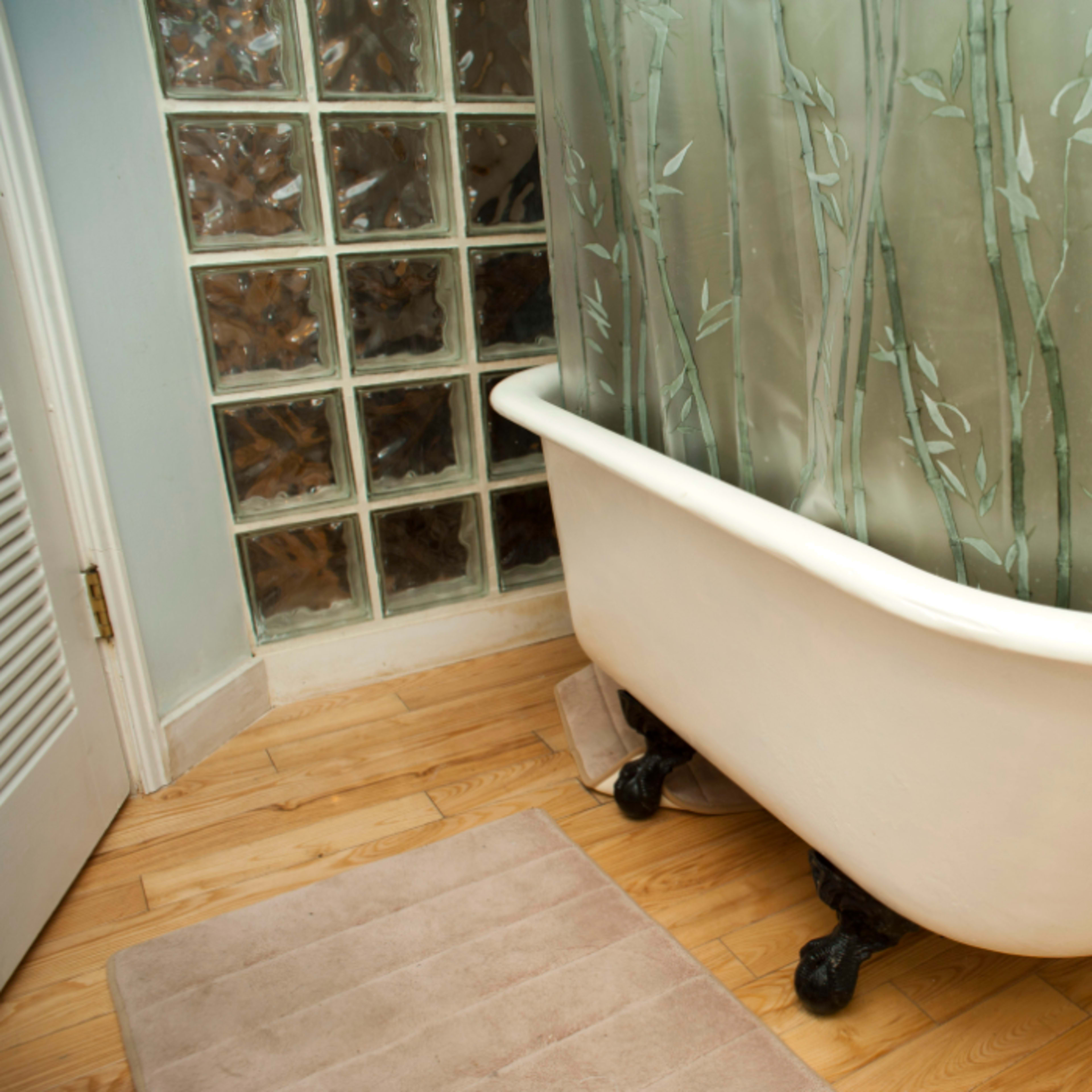 A clawfoot bathtub with a green bamboo-patterned shower curtain is positioned in a bathroom near a glass block wall.