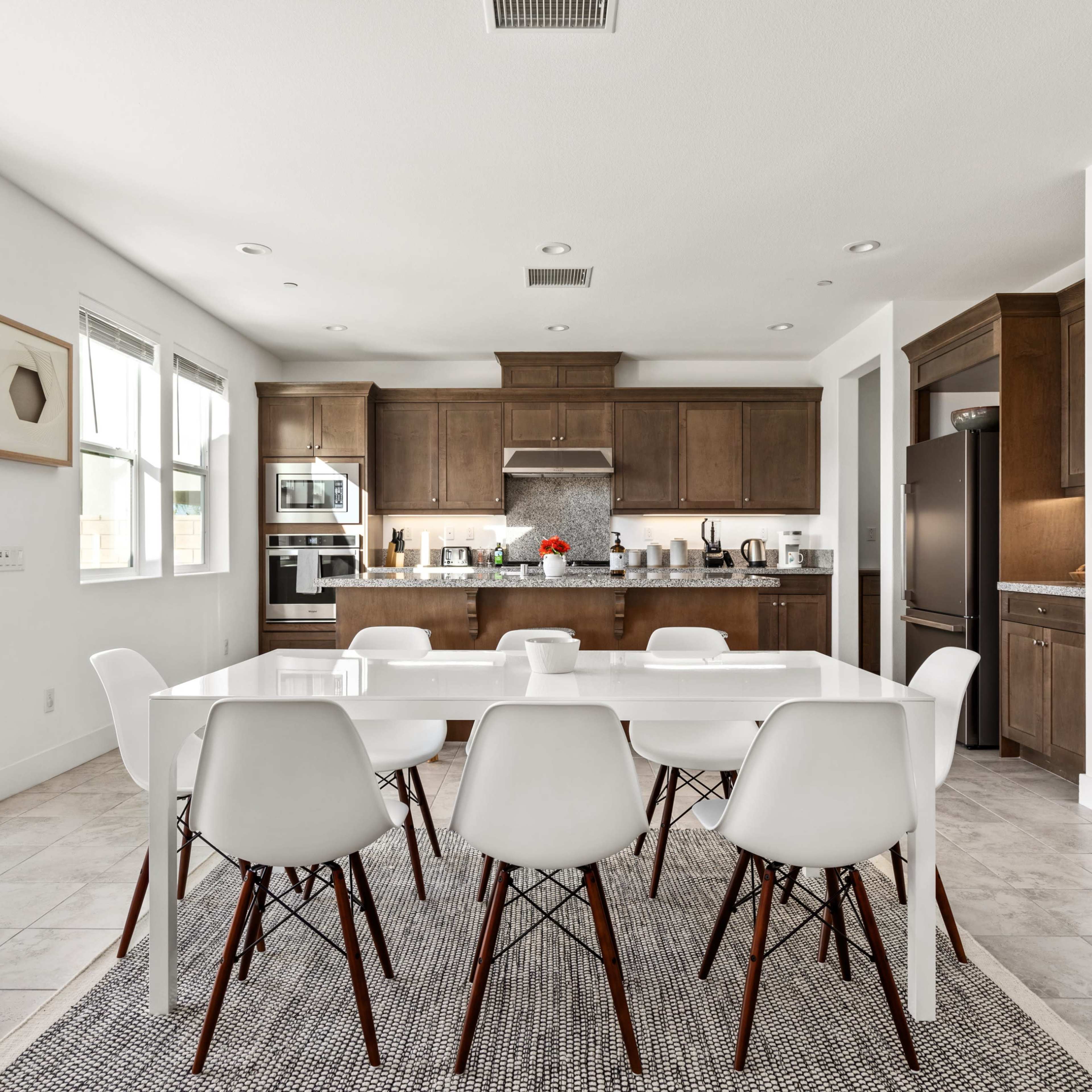 A modern kitchen features dark wooden cabinets, stainless steel appliances, and a dining table surrounded by white chairs on a patterned rug.
