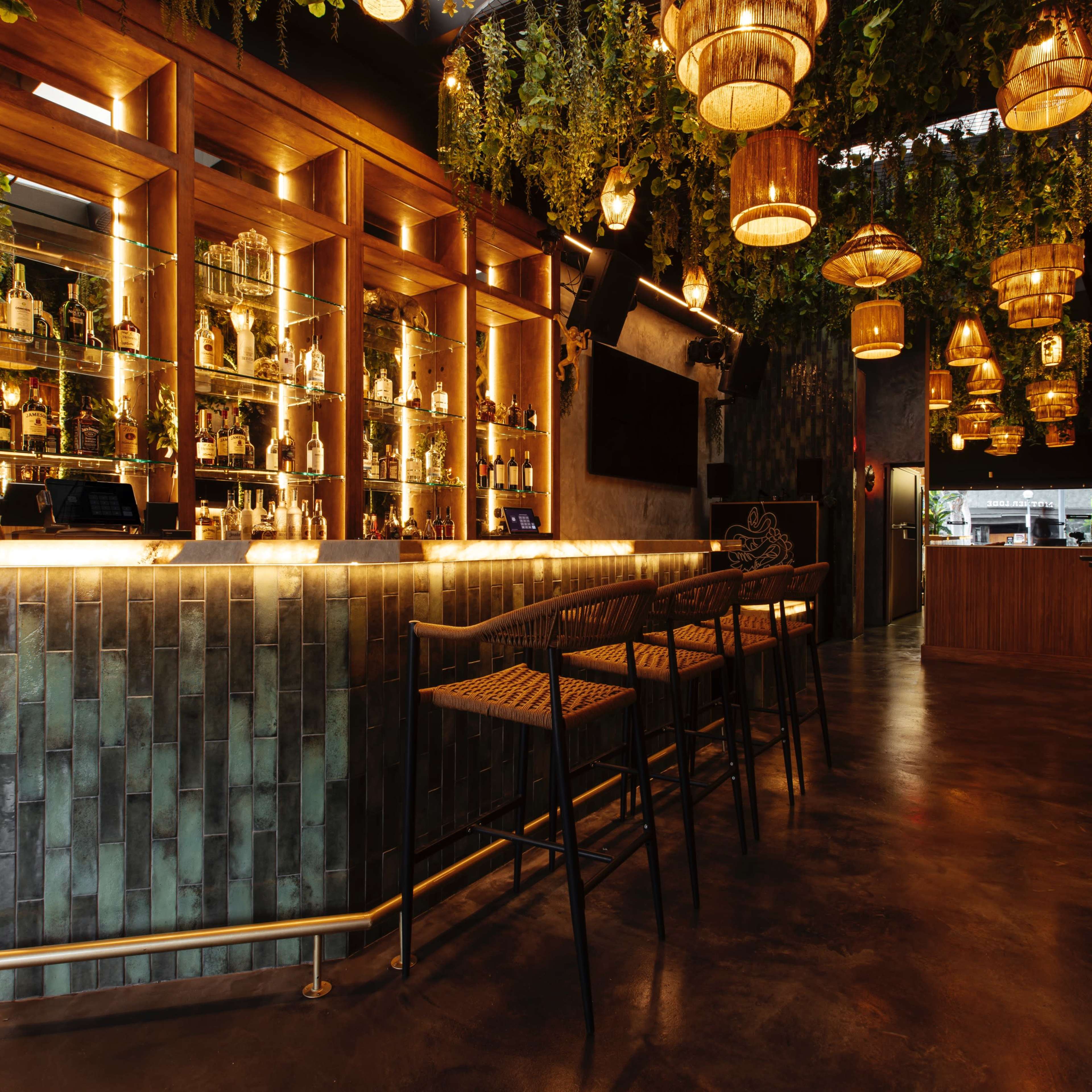 A stylish bar interior featuring a well-stocked display behind the bar, surrounded by hanging plants and lanterns, with high stools arranged in front.