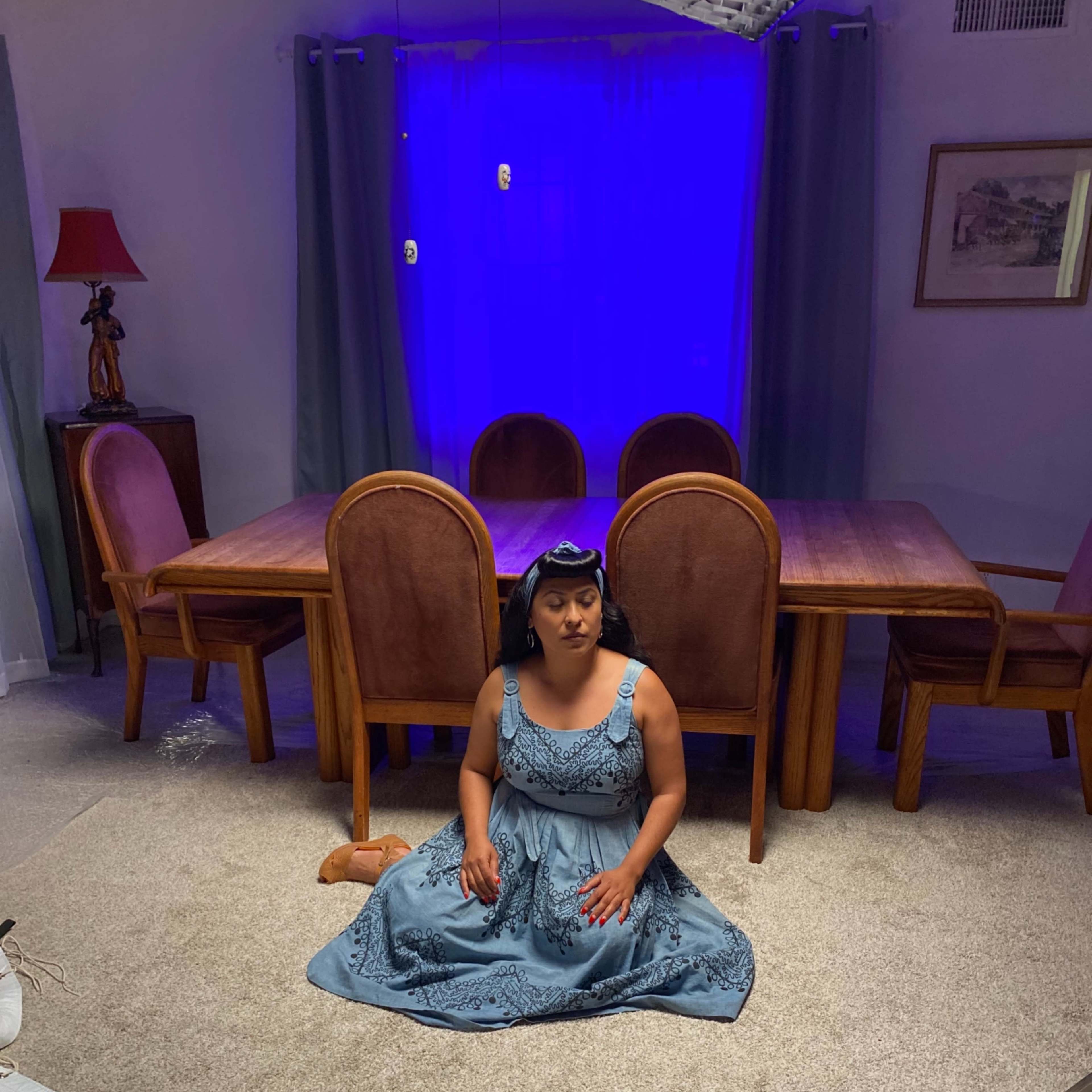 A person in a blue dress sits on the carpet in a dimly lit room with a dining table and chairs in the background.