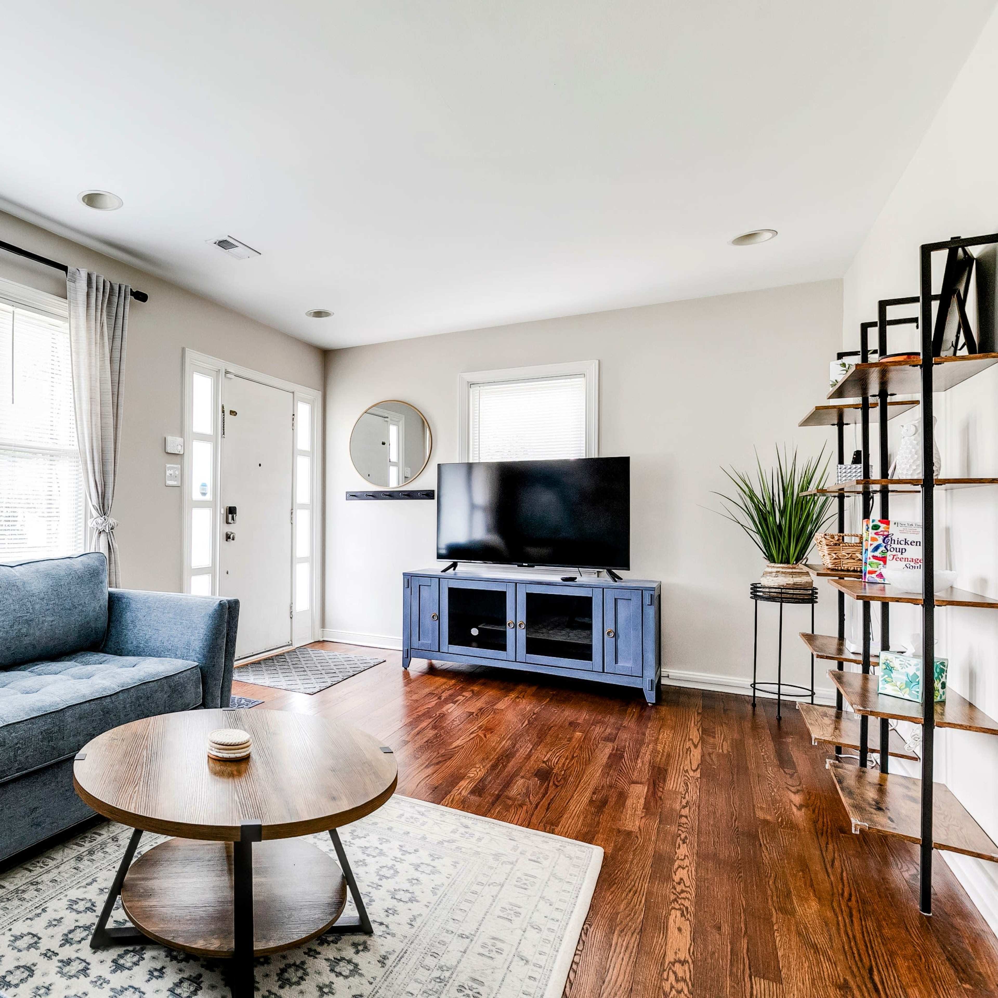 A modern living room features a blue sectional sofa, a round wooden coffee table, a television on a stand, and a mix of natural light from large windows.