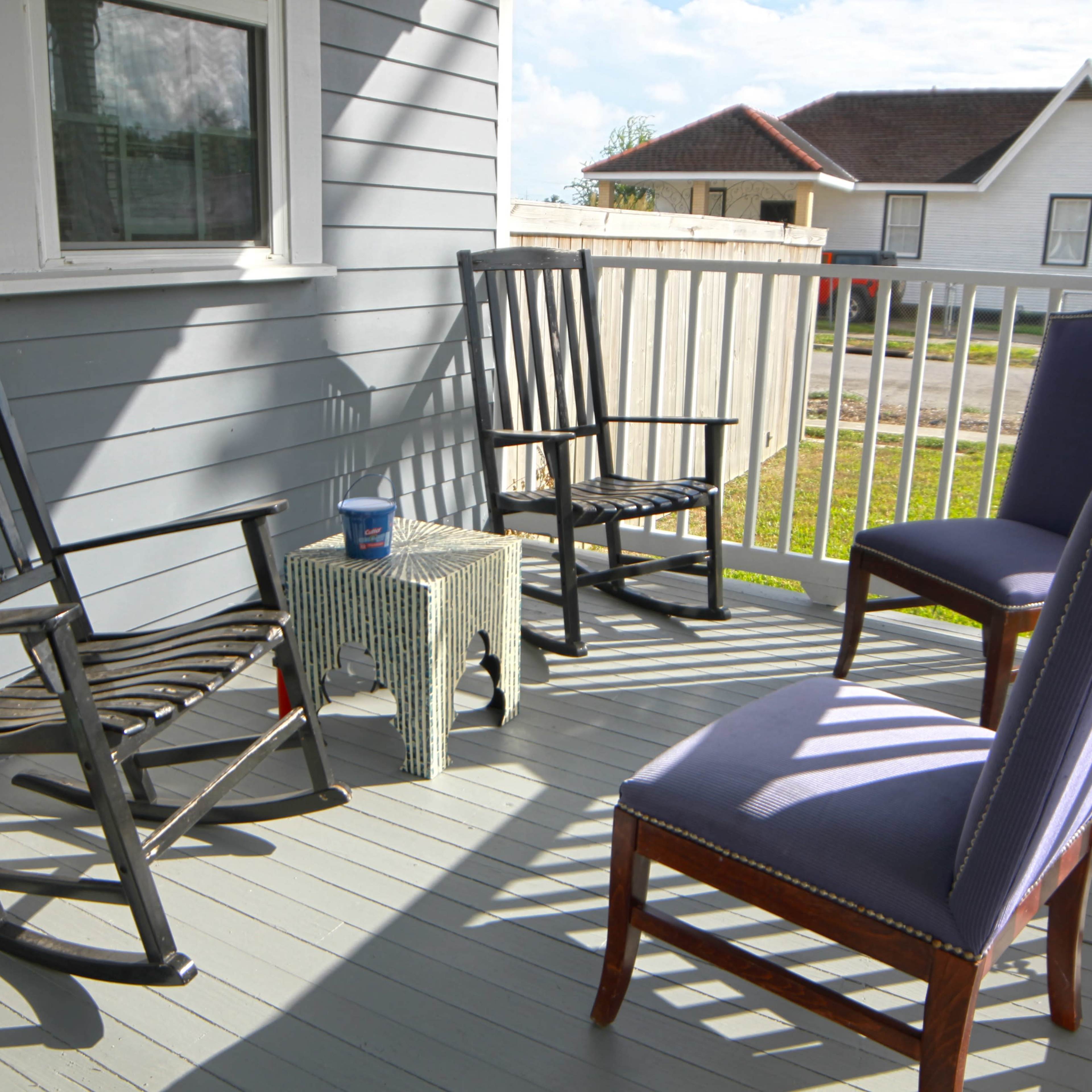 A porch with two rocking chairs, a small table, and two upright chairs against a backdrop of a nearby house and green lawn.