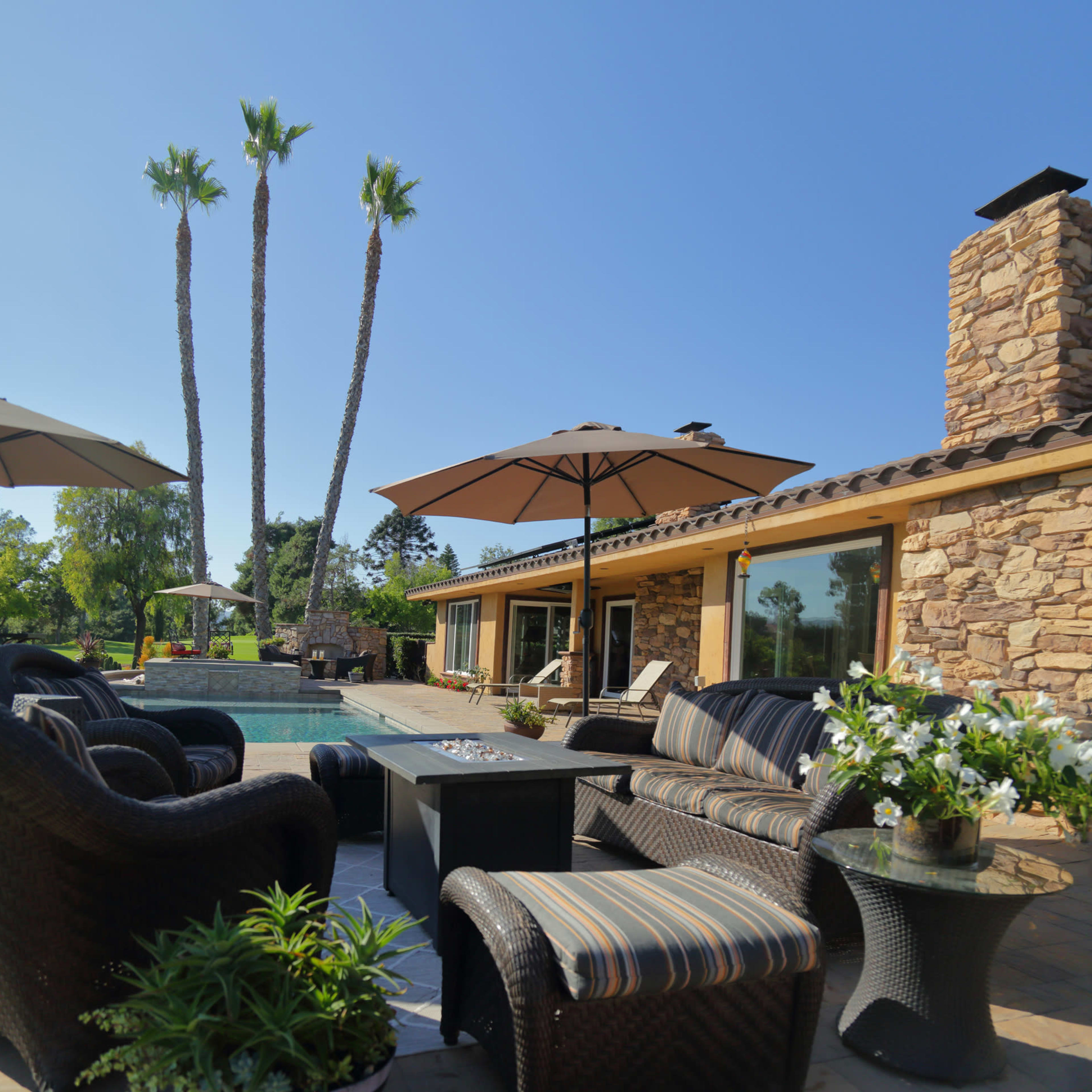 A patio area with seating and umbrellas surrounds a swimming pool, accompanied by palm trees and a stone house in the background.