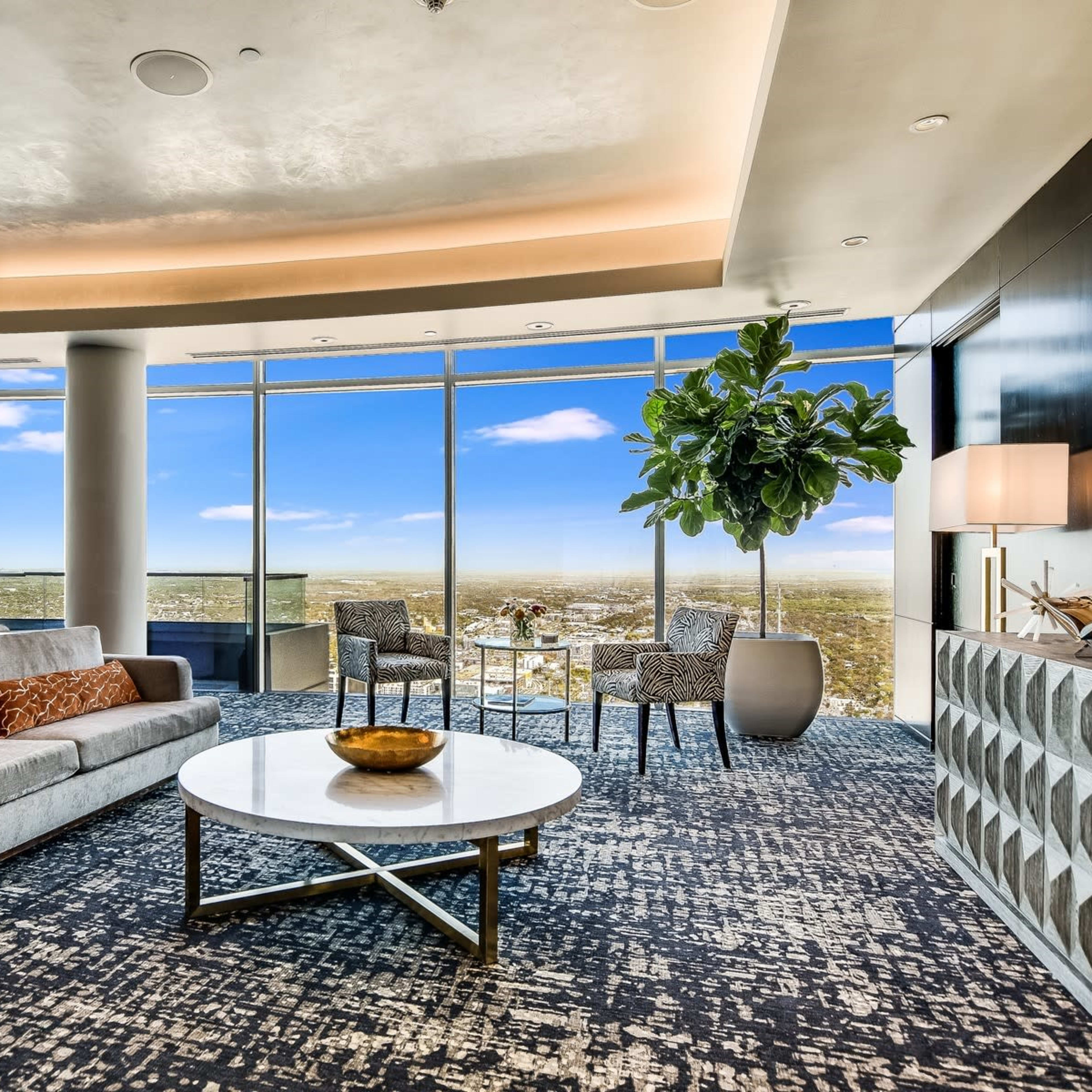 A modern living room features a gray sofa with decorative pillows, a round coffee table, two chairs, and large windows showcasing a panoramic view of the landscape.