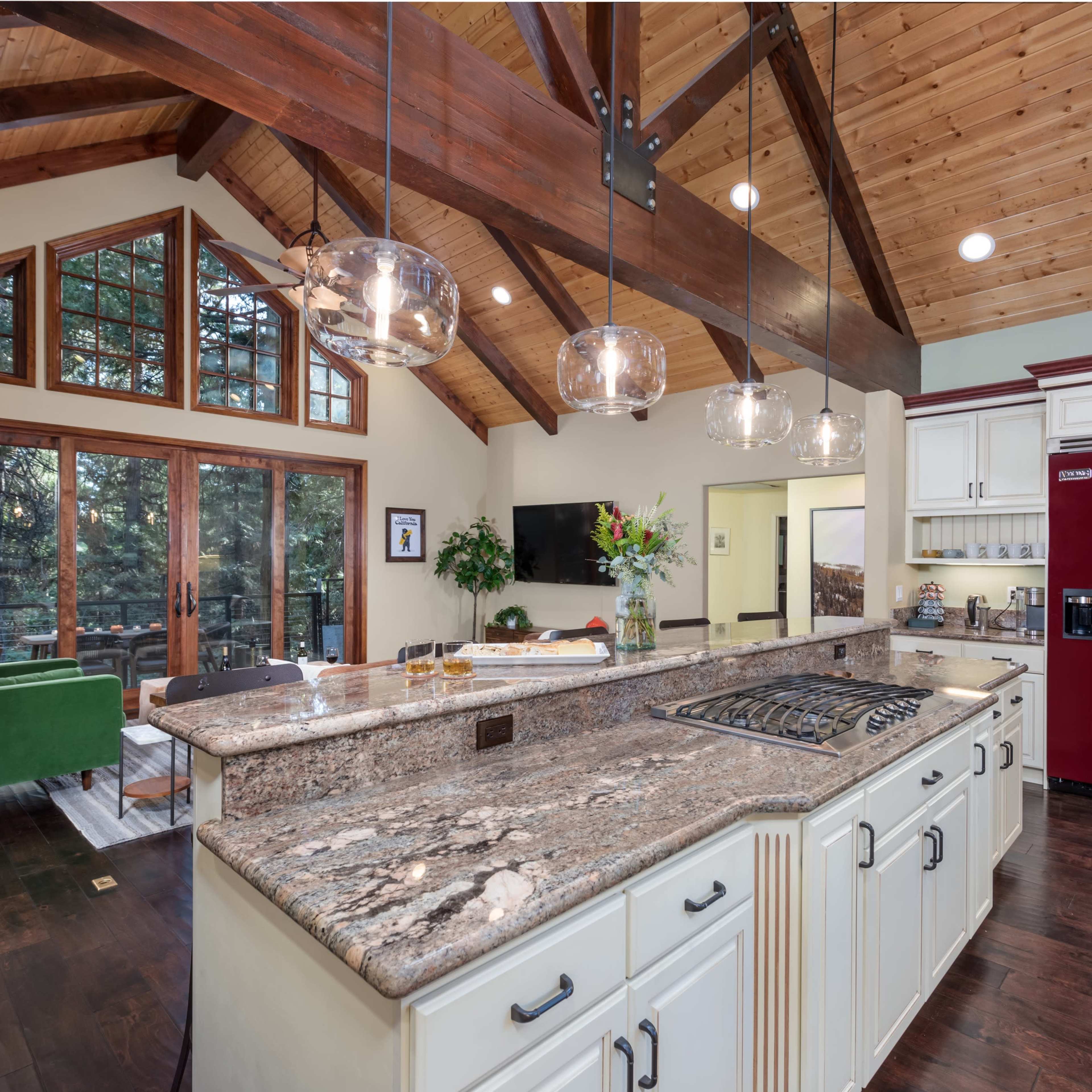 A spacious kitchen features a large granite island, modern appliances, and vaulted wooden beams, with large windows providing views of the outdoors.