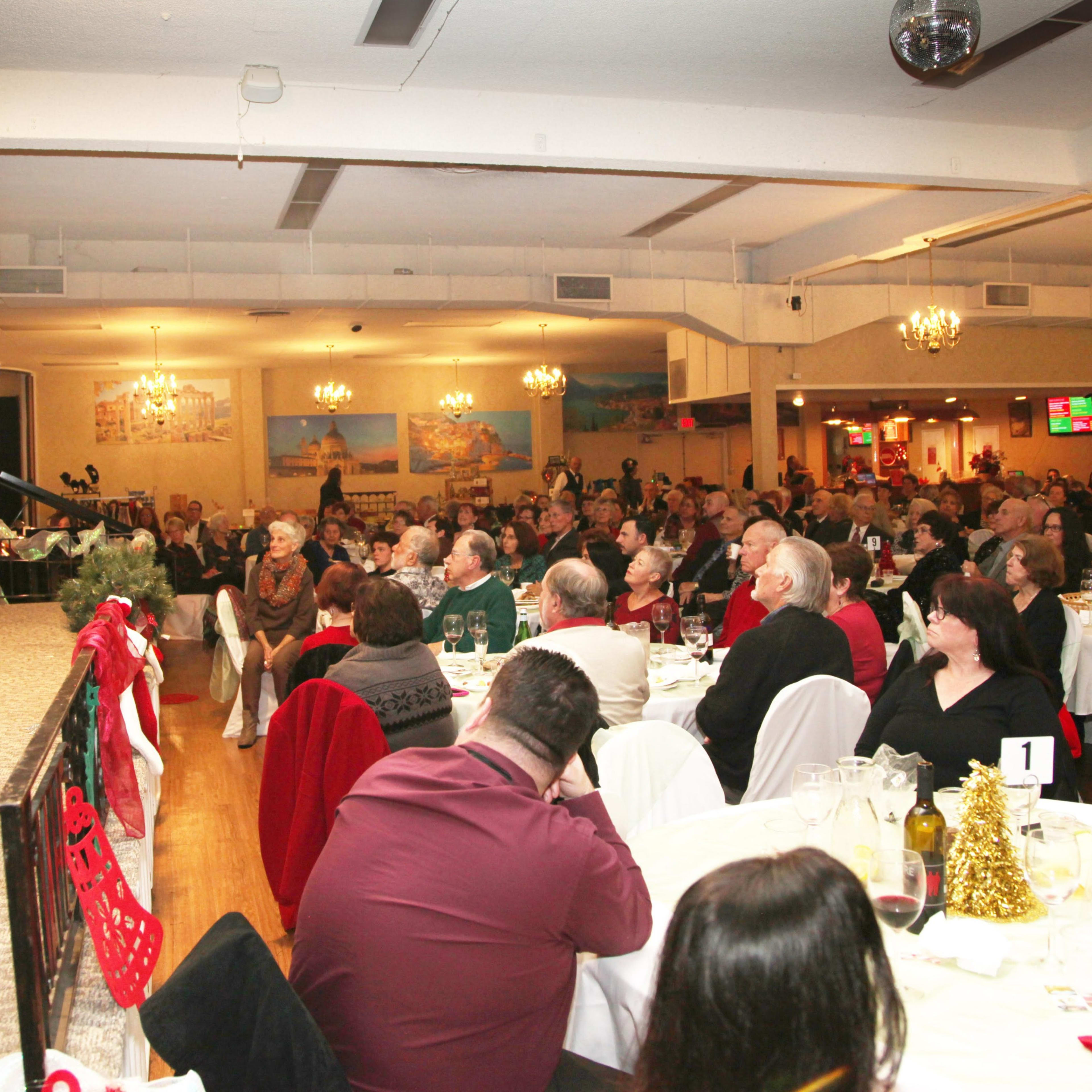 A woman stands on a stage addressing a large audience seated at tables adorned with festive decorations.
