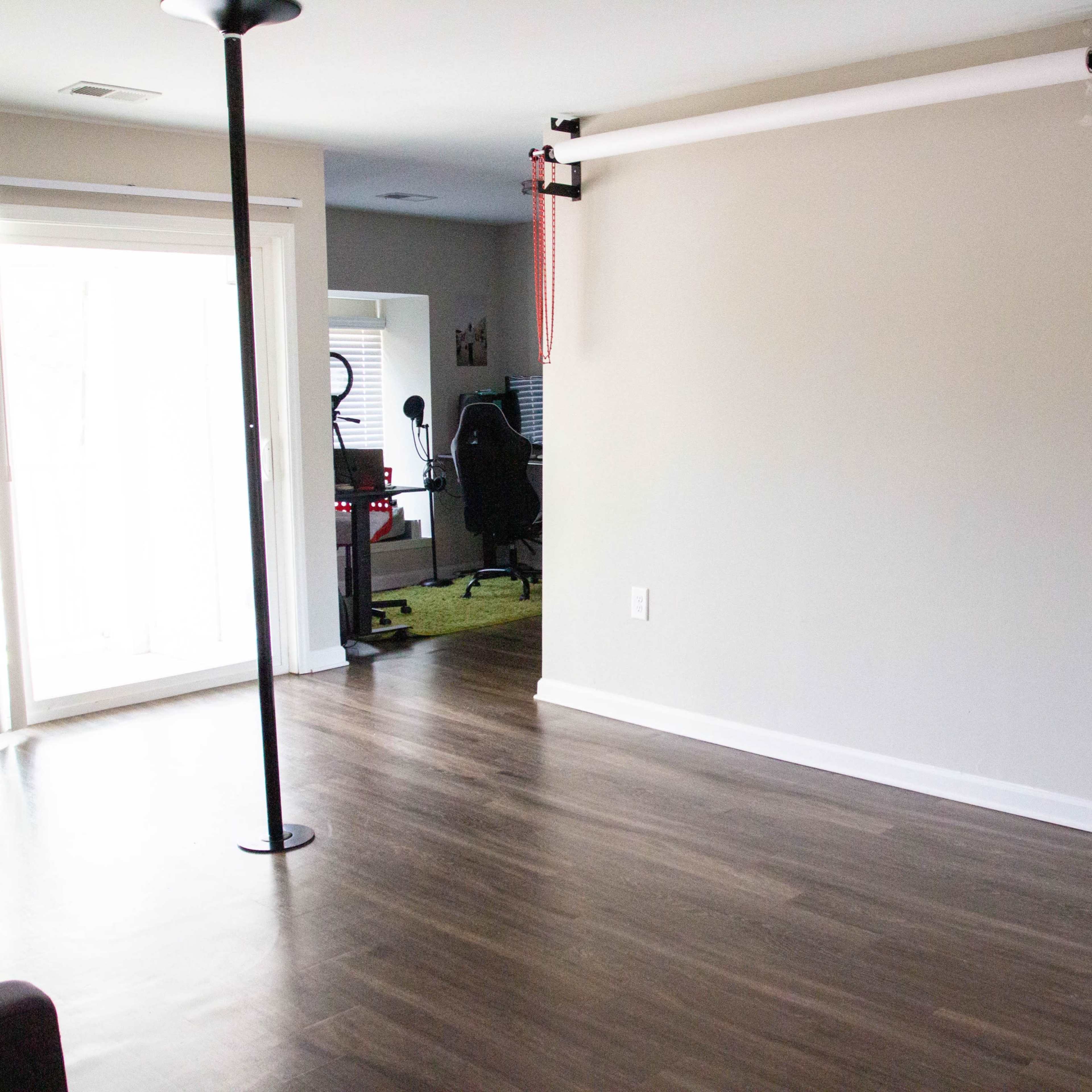 A spacious room features hardwood flooring, a ceiling-mounted curtain rod, and sliding glass doors leading to an outdoor area, with a desk and gaming chair visible in the background.