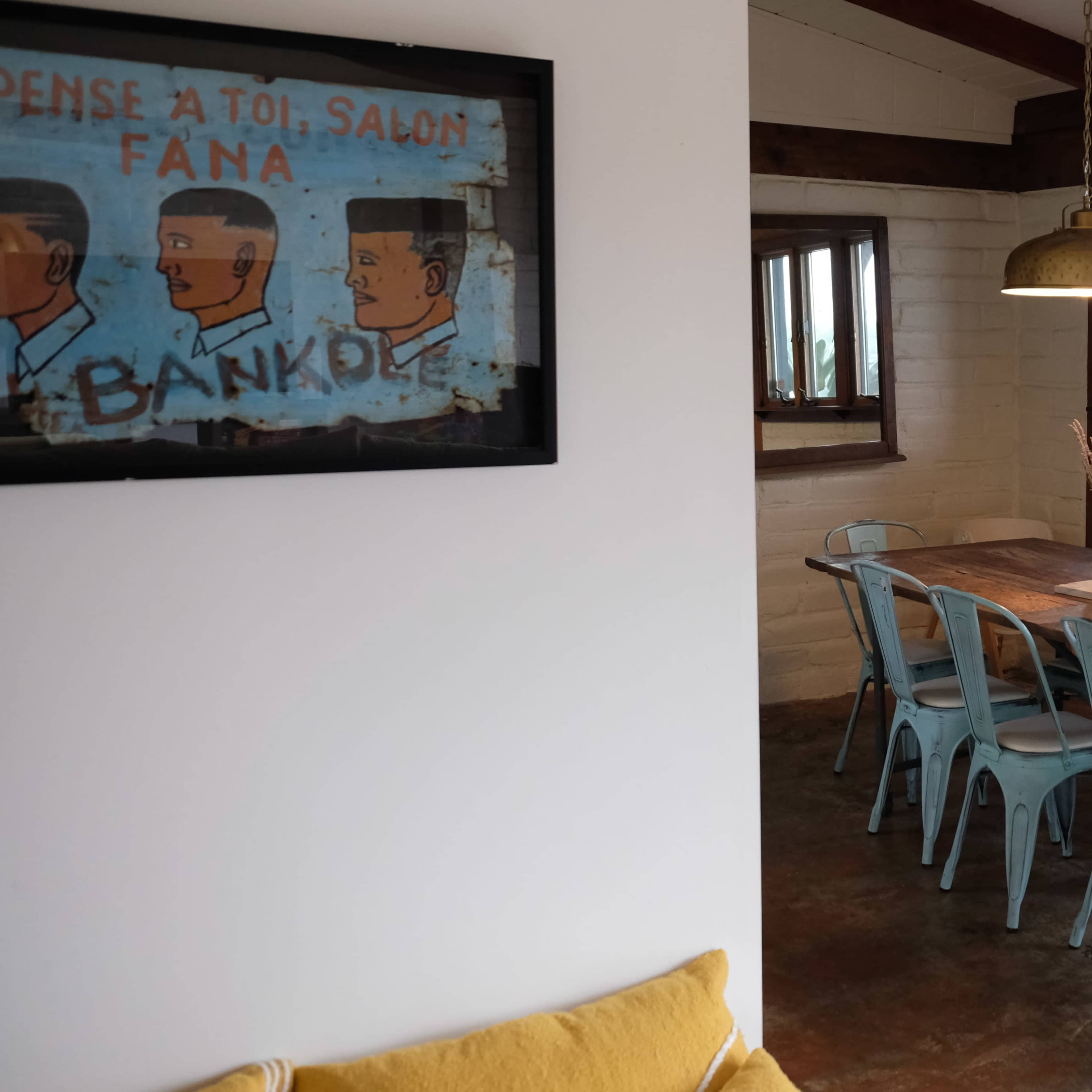 A rustic dining area featuring a wooden table surrounded by blue metal chairs, with large windows and a pendant light overhead.