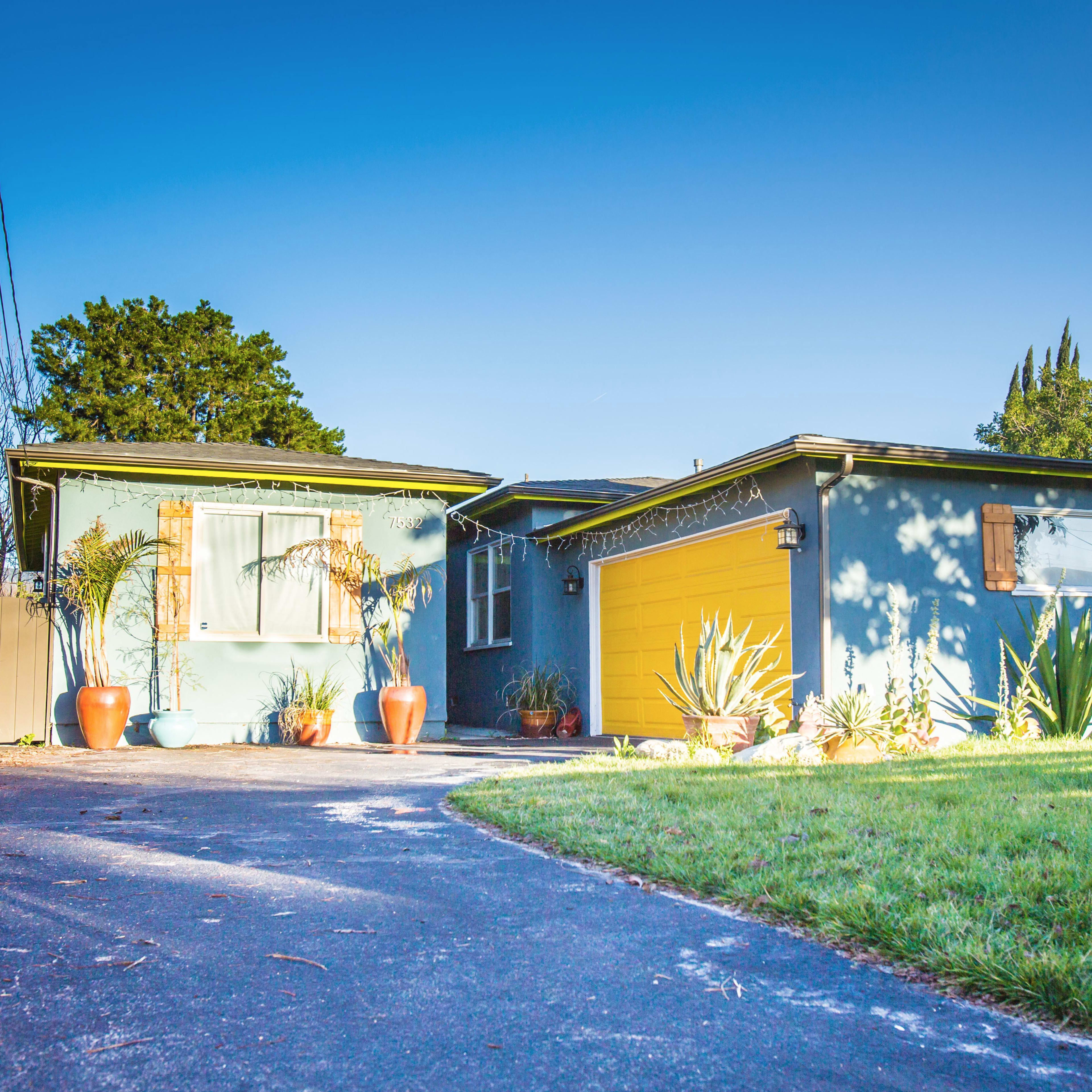 A blue house with a yellow garage door, surrounded by potted plants and a driveway leading up to it.