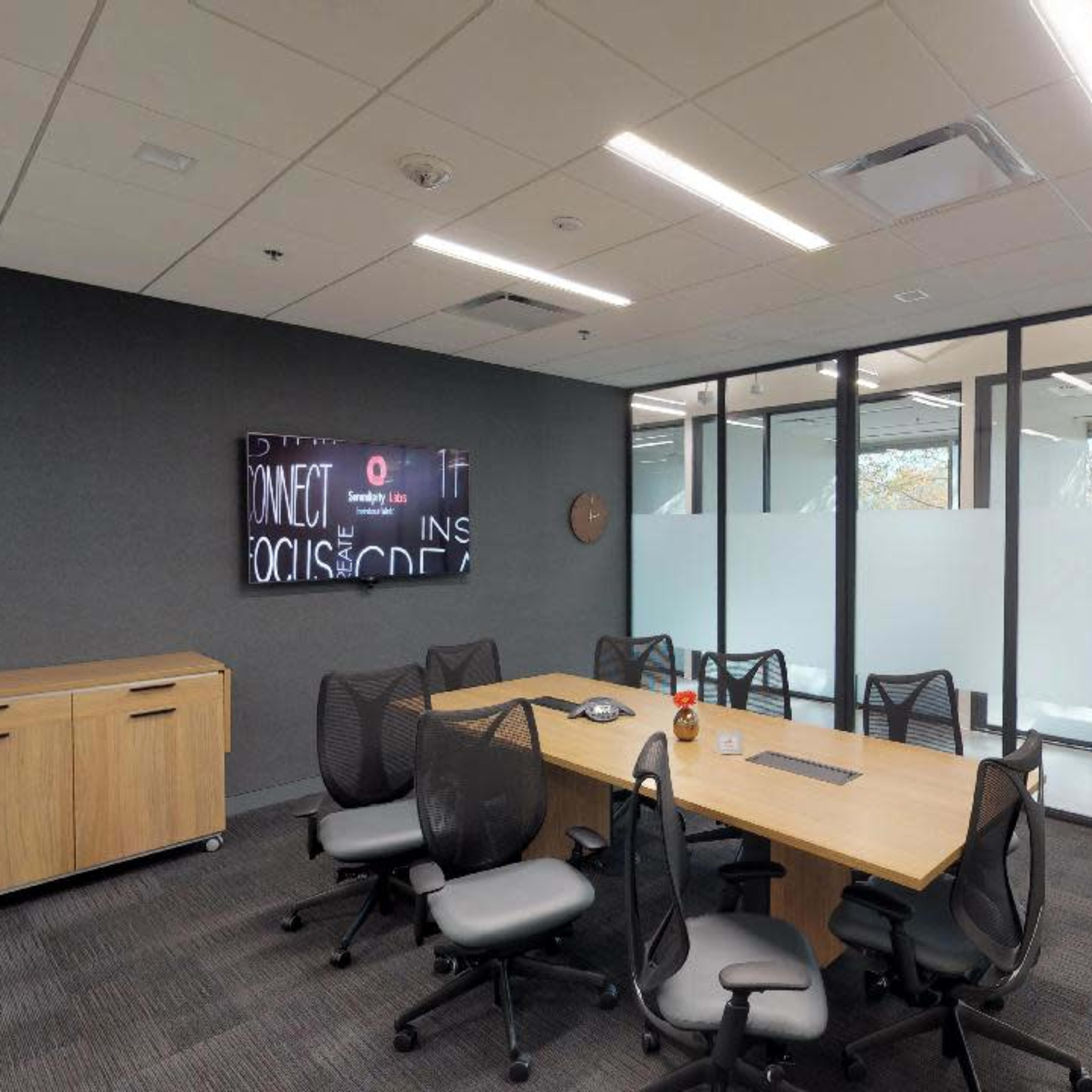 A modern conference room features a large wooden table surrounded by ergonomic chairs, with a wall-mounted screen and glass partitions.