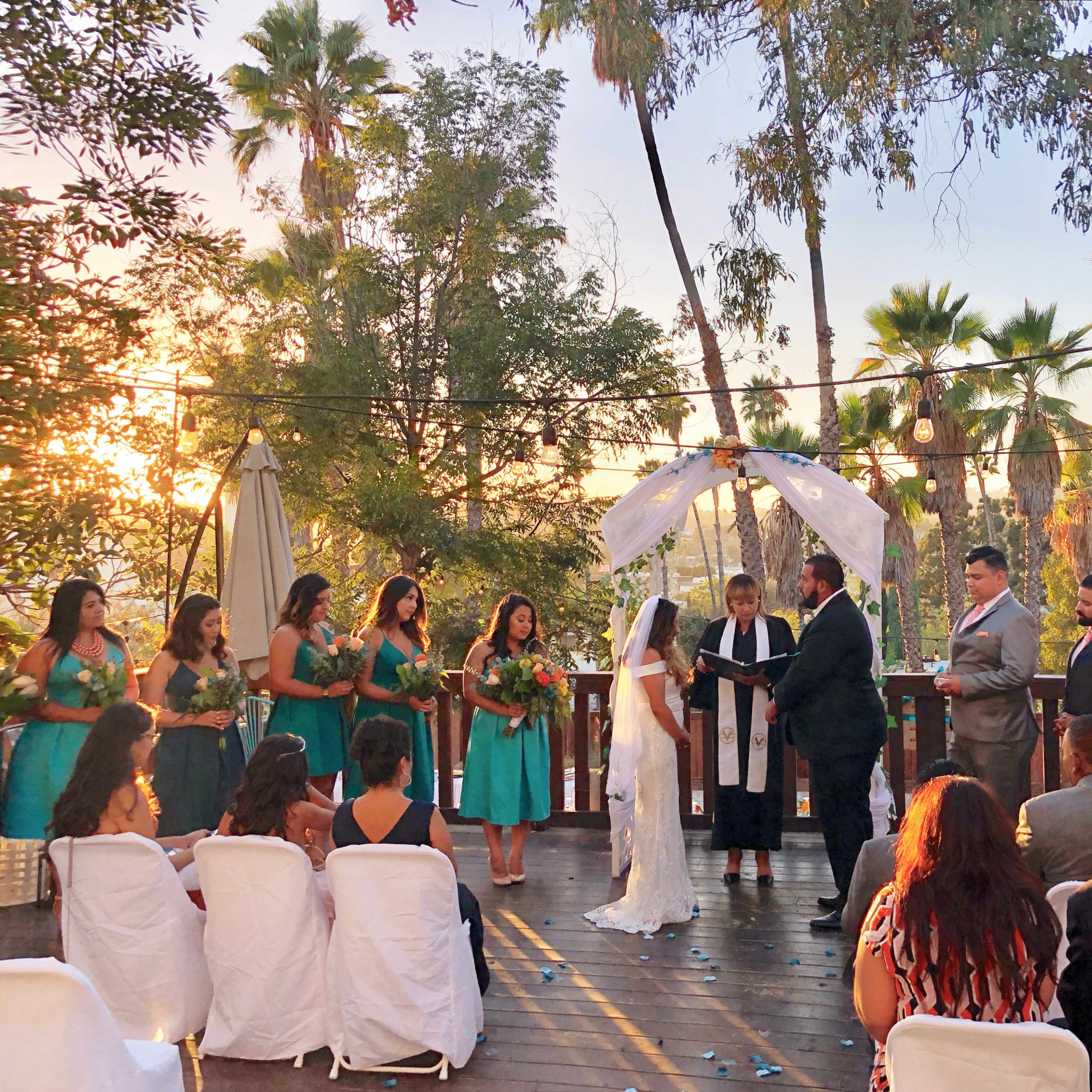 A wedding ceremony is taking place outdoors on a deck surrounded by palm trees, with the bride and groom standing under a floral arch.
