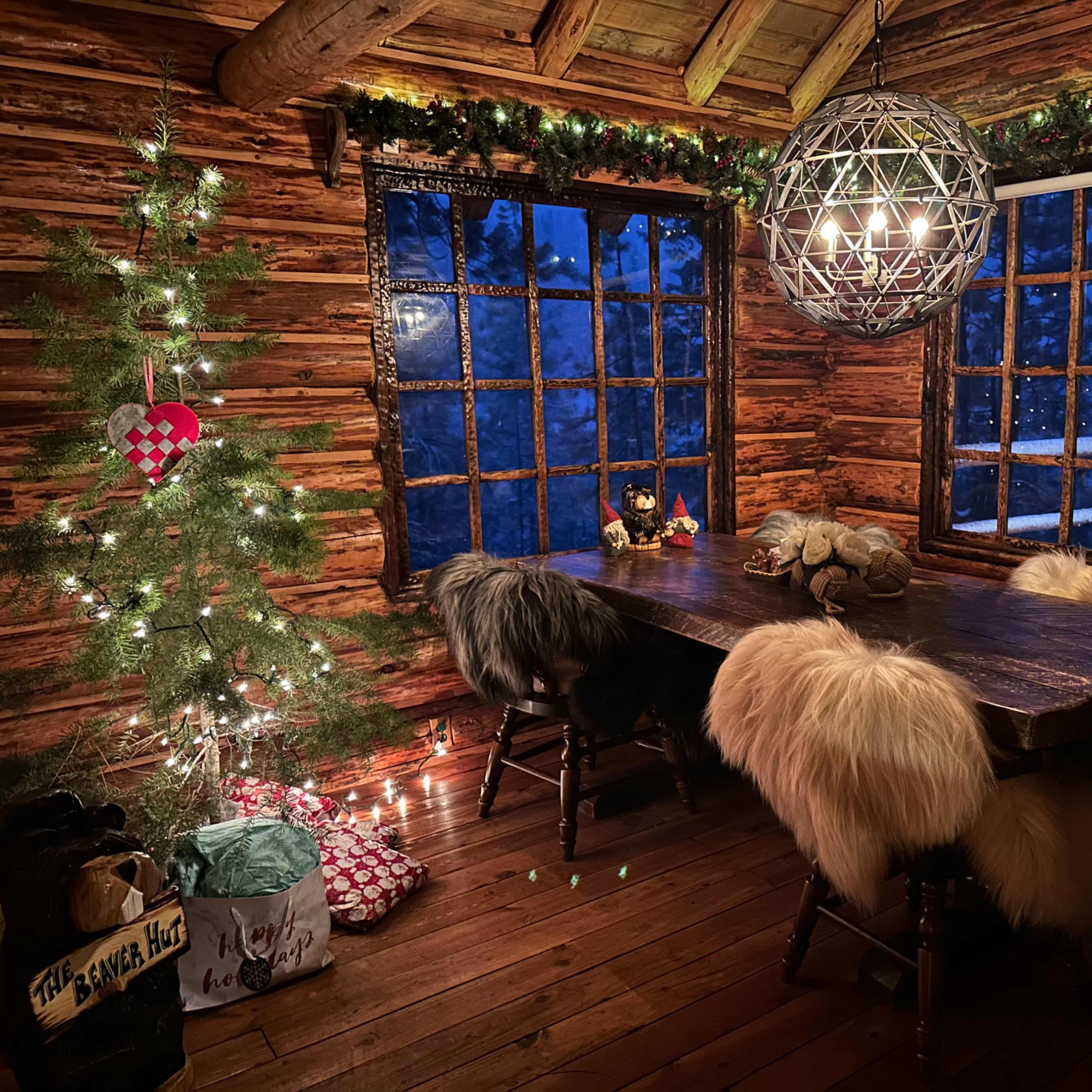 A rustic wooden dining area features a large table surrounded by fur-covered chairs, with a Christmas tree and decorations beside the windows.