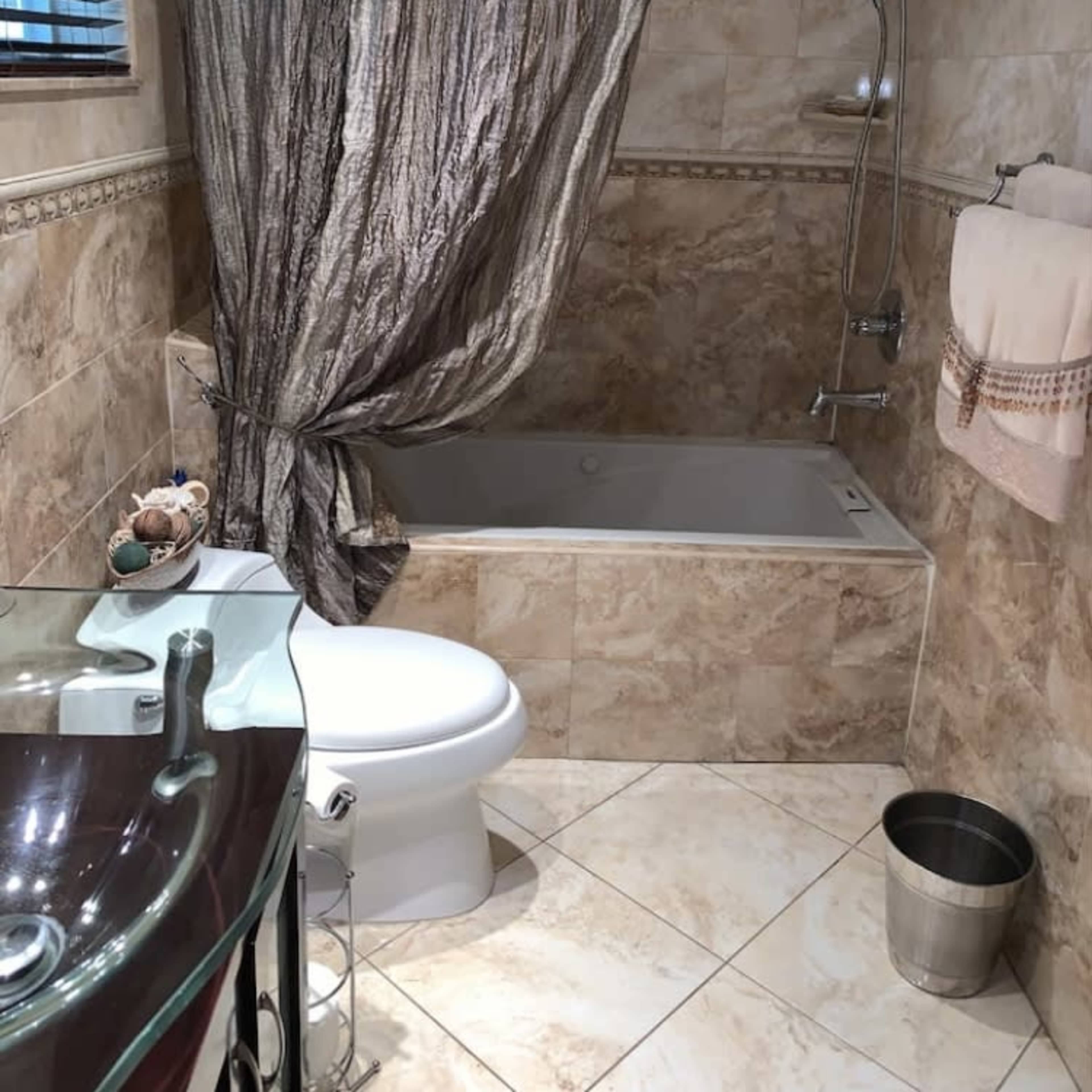 A bathroom with a bathtub, shower, toilet, a glass sink, and a decorative curtain.