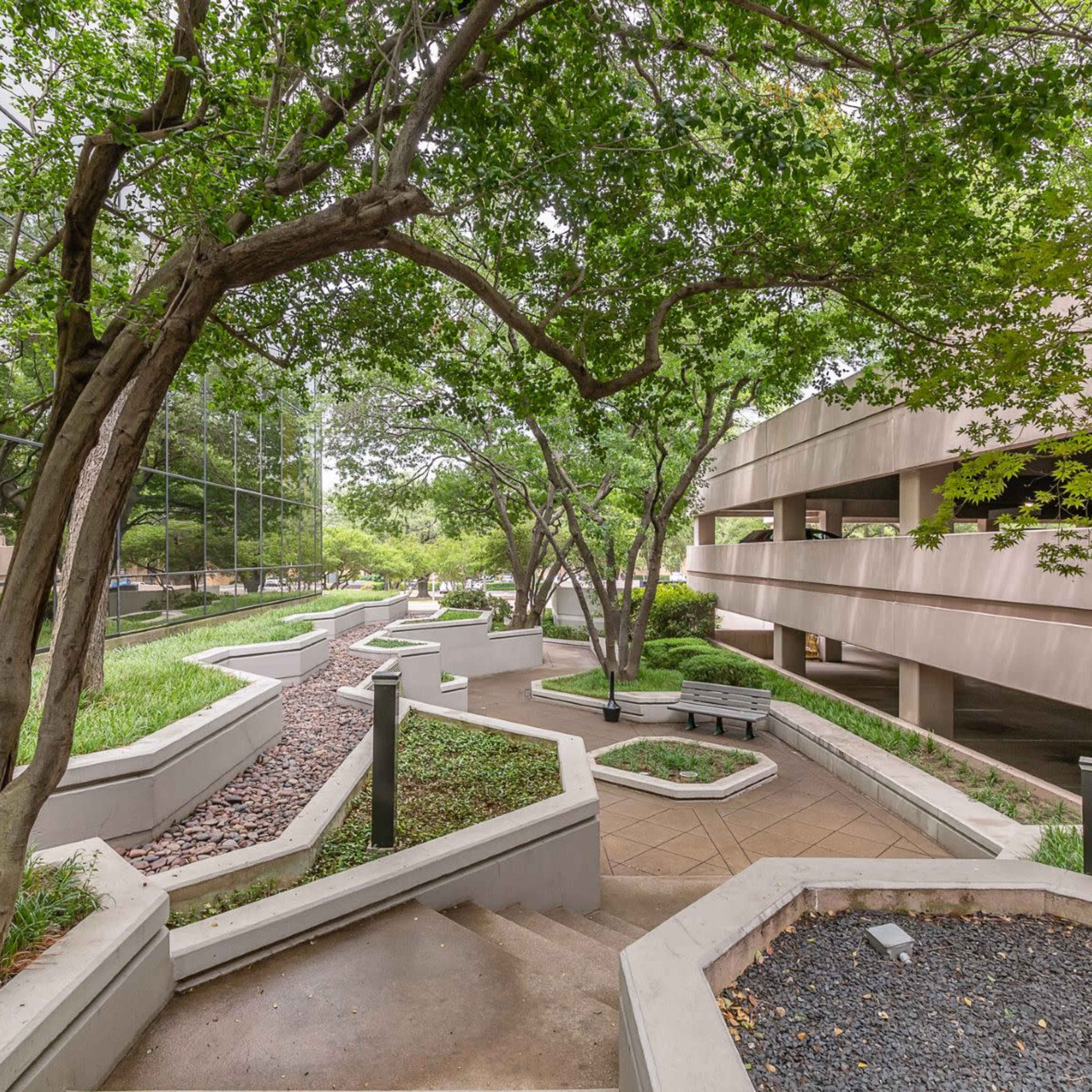 A landscaped walkway with curved paths, surrounded by trees and a reflective building, leading to a parking garage.