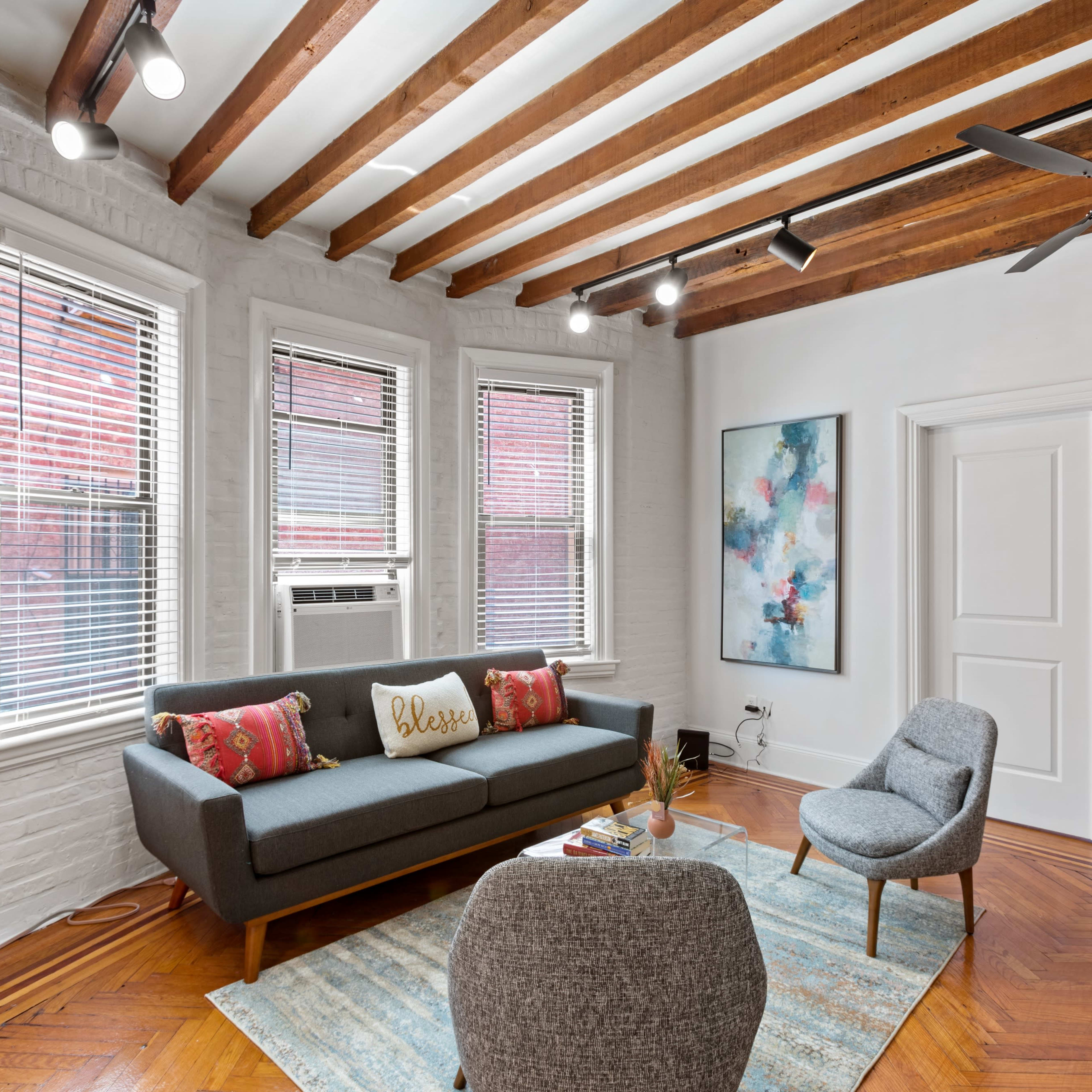 A modern living room with a grey sofa, two armchairs, hardwood floors, and exposed wooden beams on the ceiling.