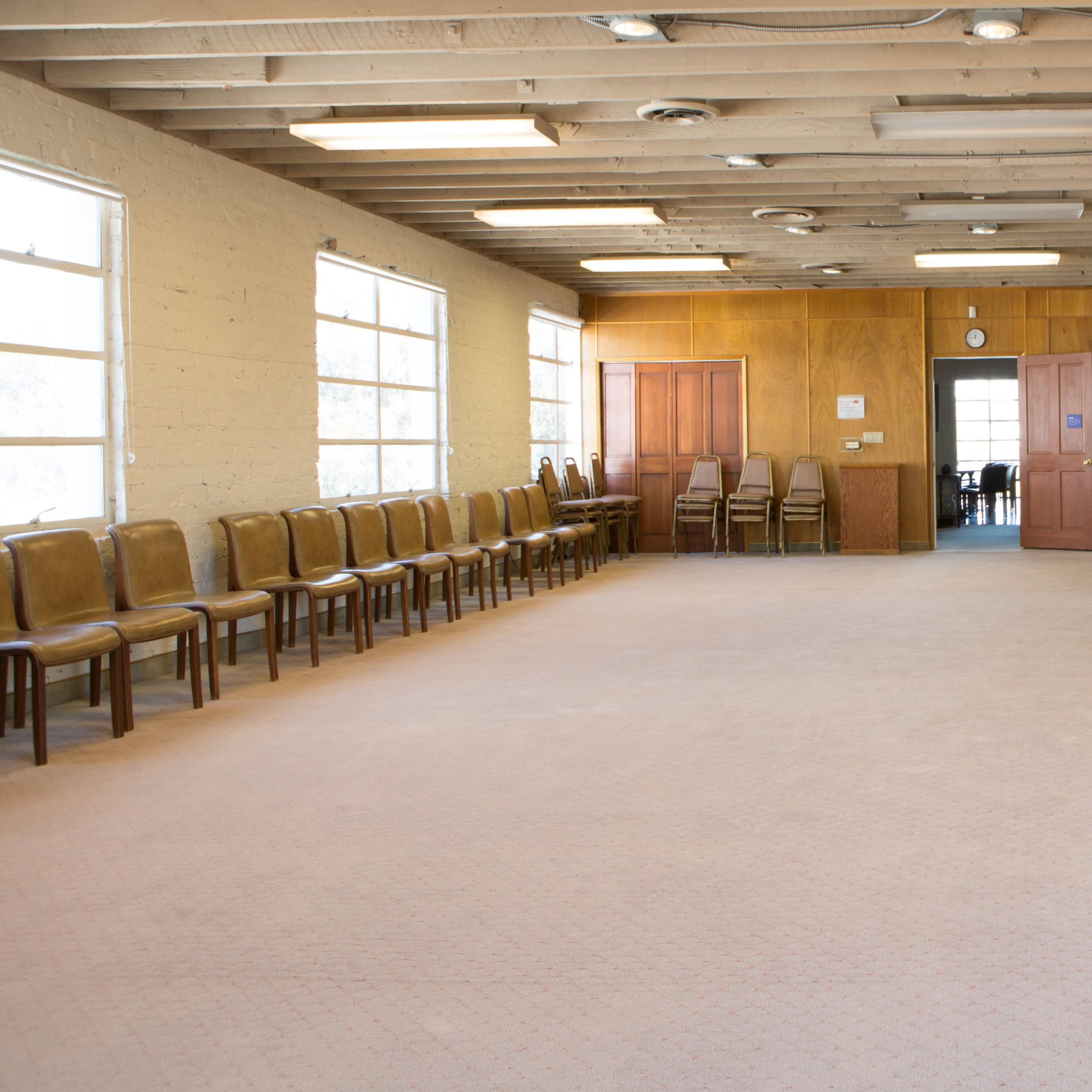 A spacious, empty room with wooden paneling, arranged seating along the walls, and a piano in one corner.