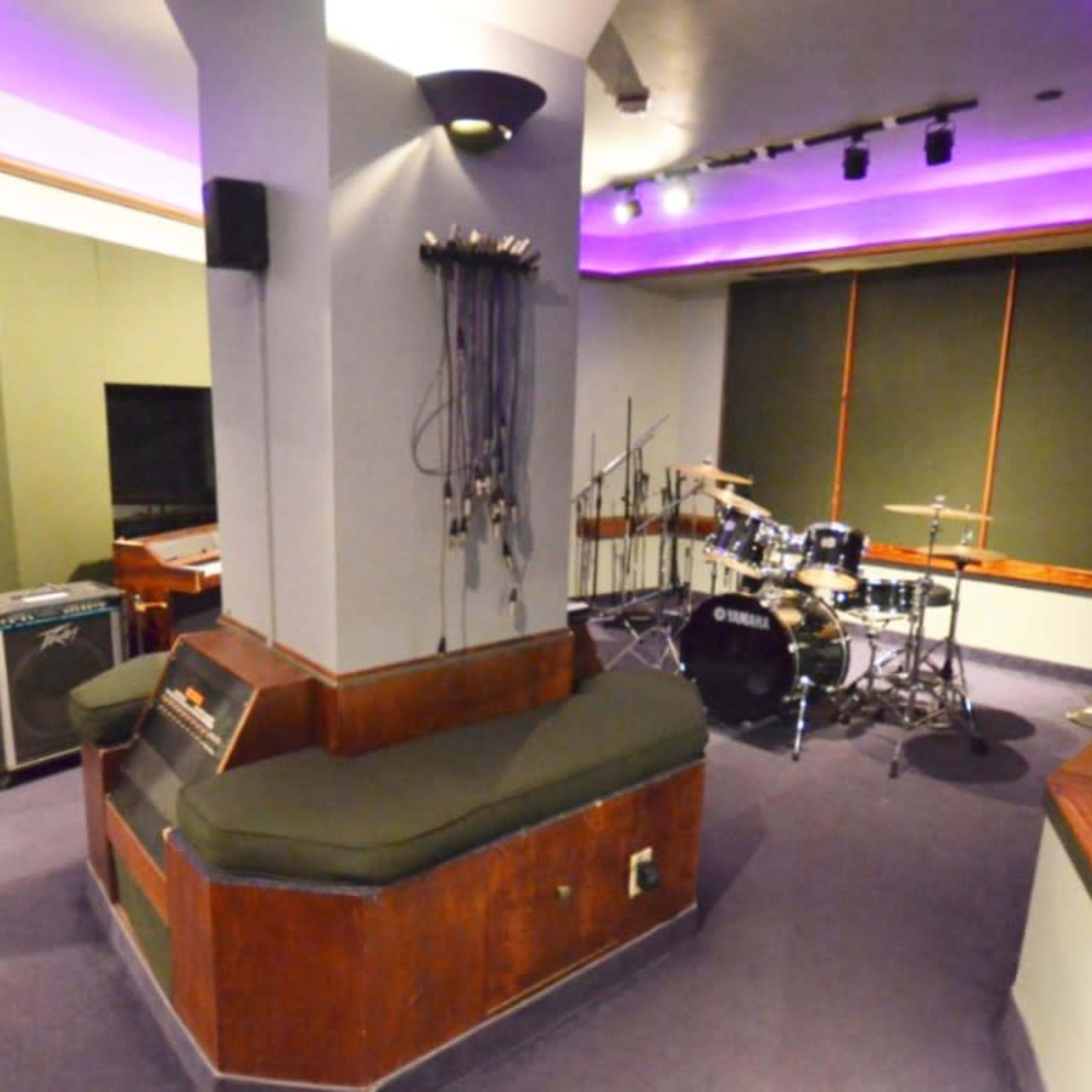 A partially furnished recording studio with musical instruments, including a keyboard, drum set, and seating area, all under purple ambient lighting.