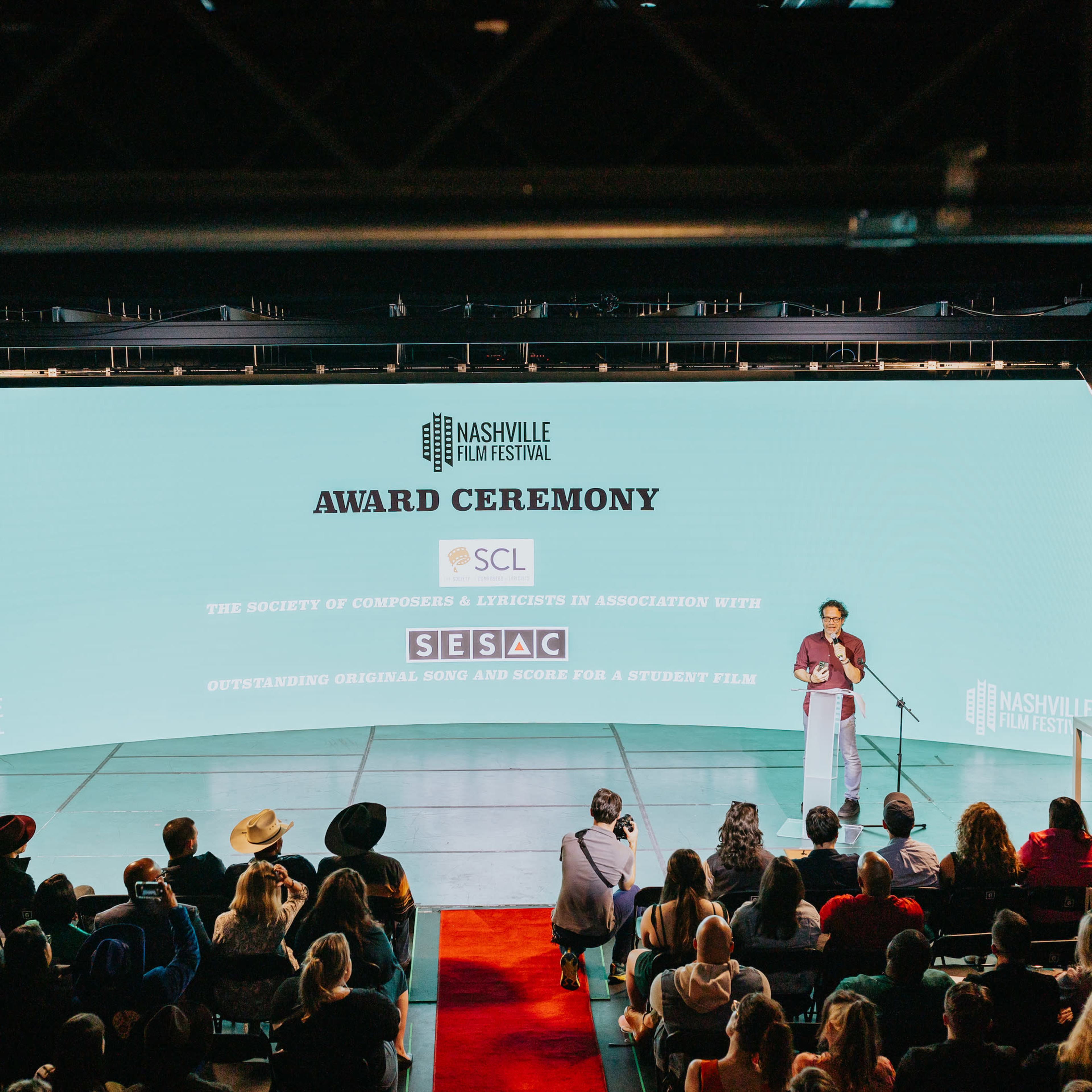 A speaker delivers a presentation at an award ceremony with an audience seated in front of a large screen displaying event details.