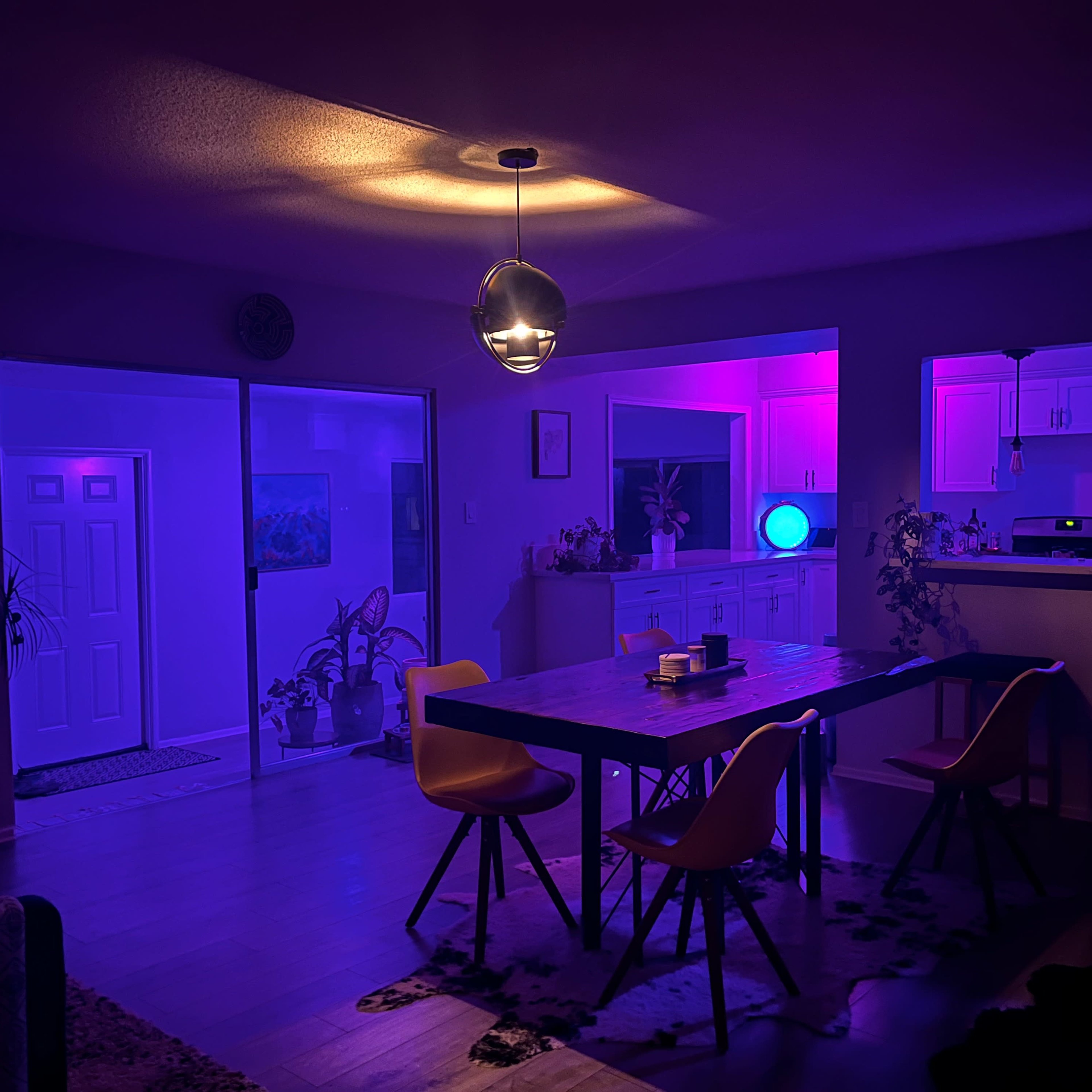A dimly lit dining area with a large table surrounded by colorful purple and blue lighting, highlighting the kitchen and entryway in the background.