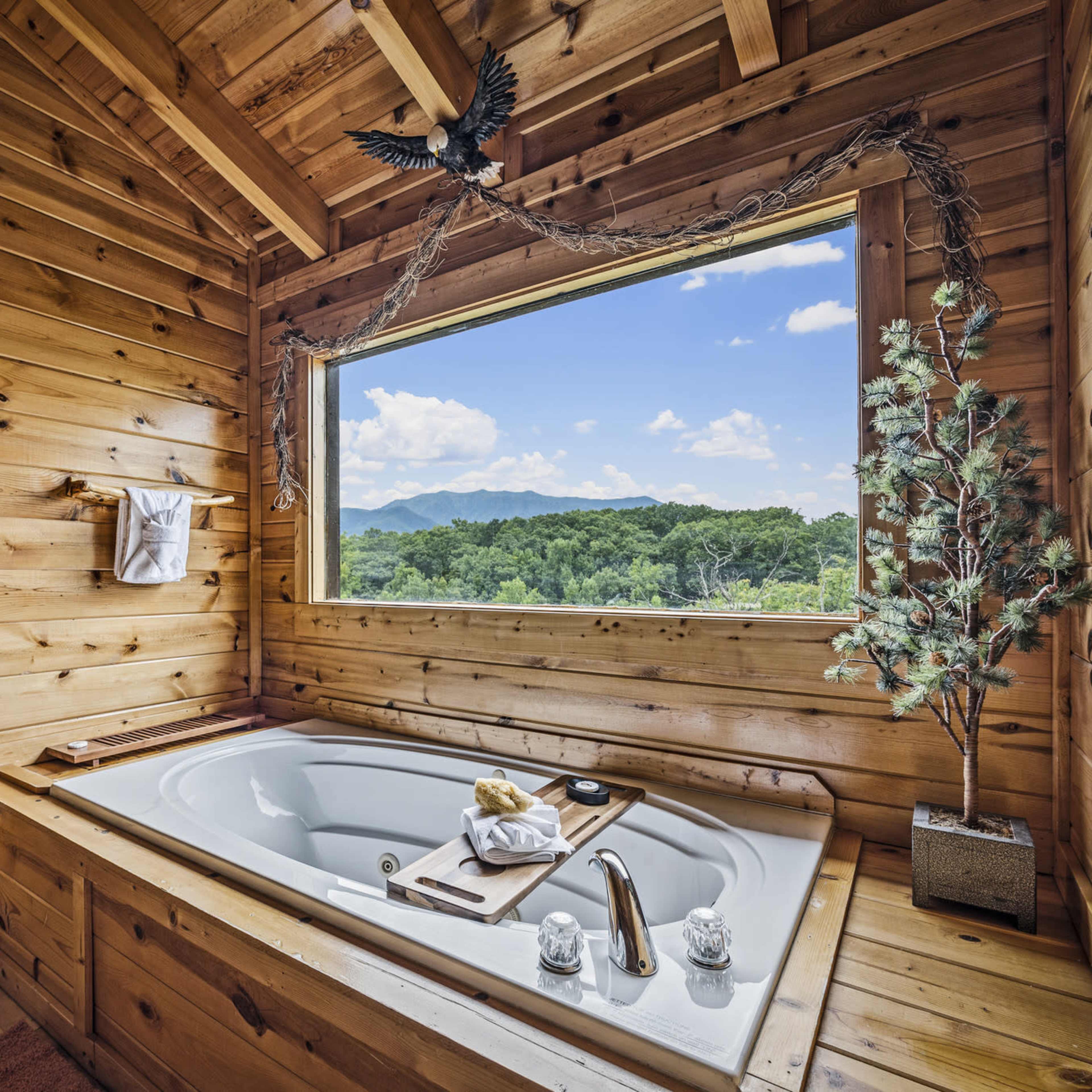 A wooden bathroom features a jetted tub positioned by a large window, offering a view of the mountains and trees outside.