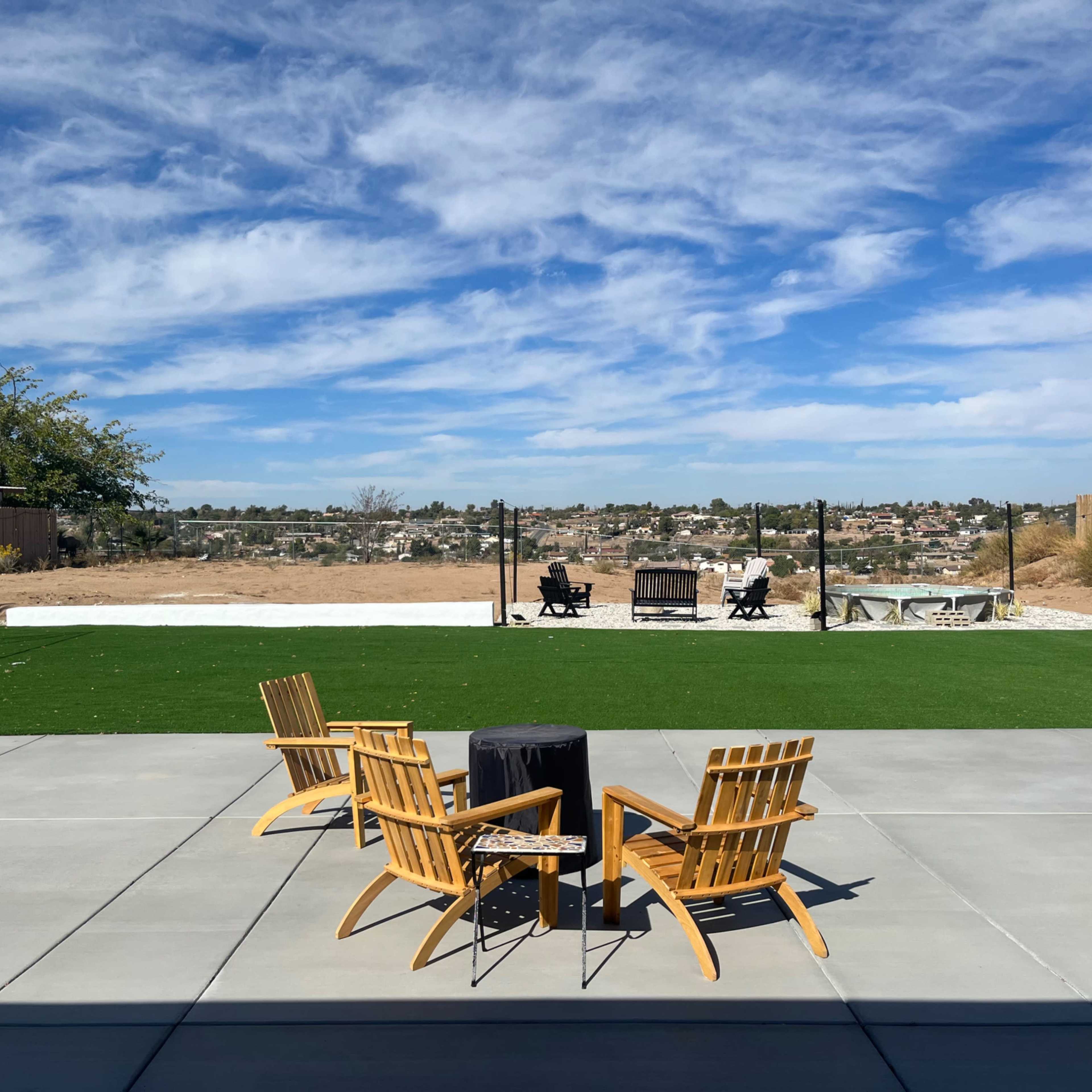 A backyard with four wooden chairs arranged around a small black table, overlooking a grassy area with two soccer goals and a view of the landscape under a blue sky with clouds.